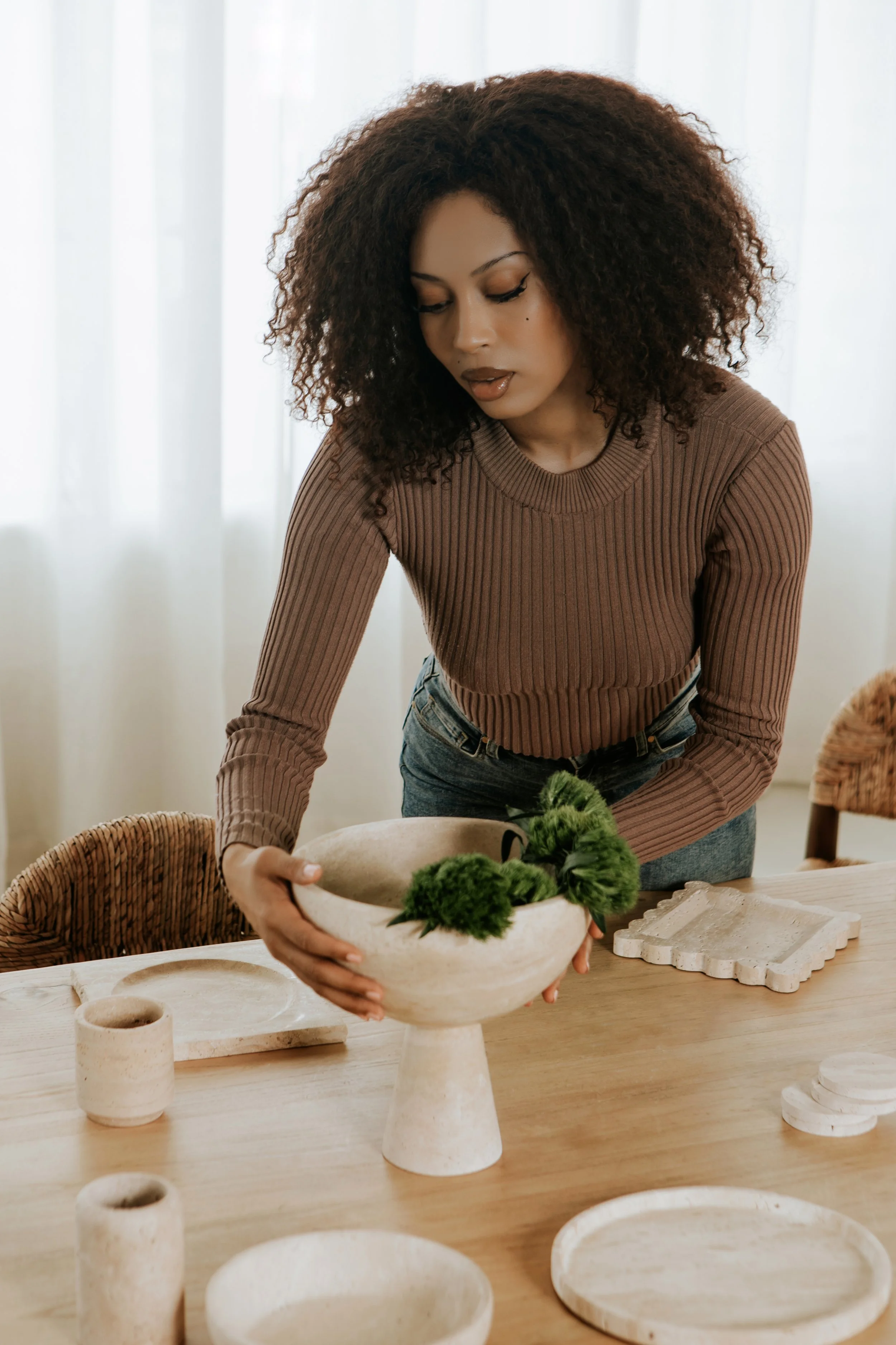 A woman with curly hair placing a ceramic bowl with green moss on a wooden table surrounded by various other ceramic items, in a bright room with white curtains.