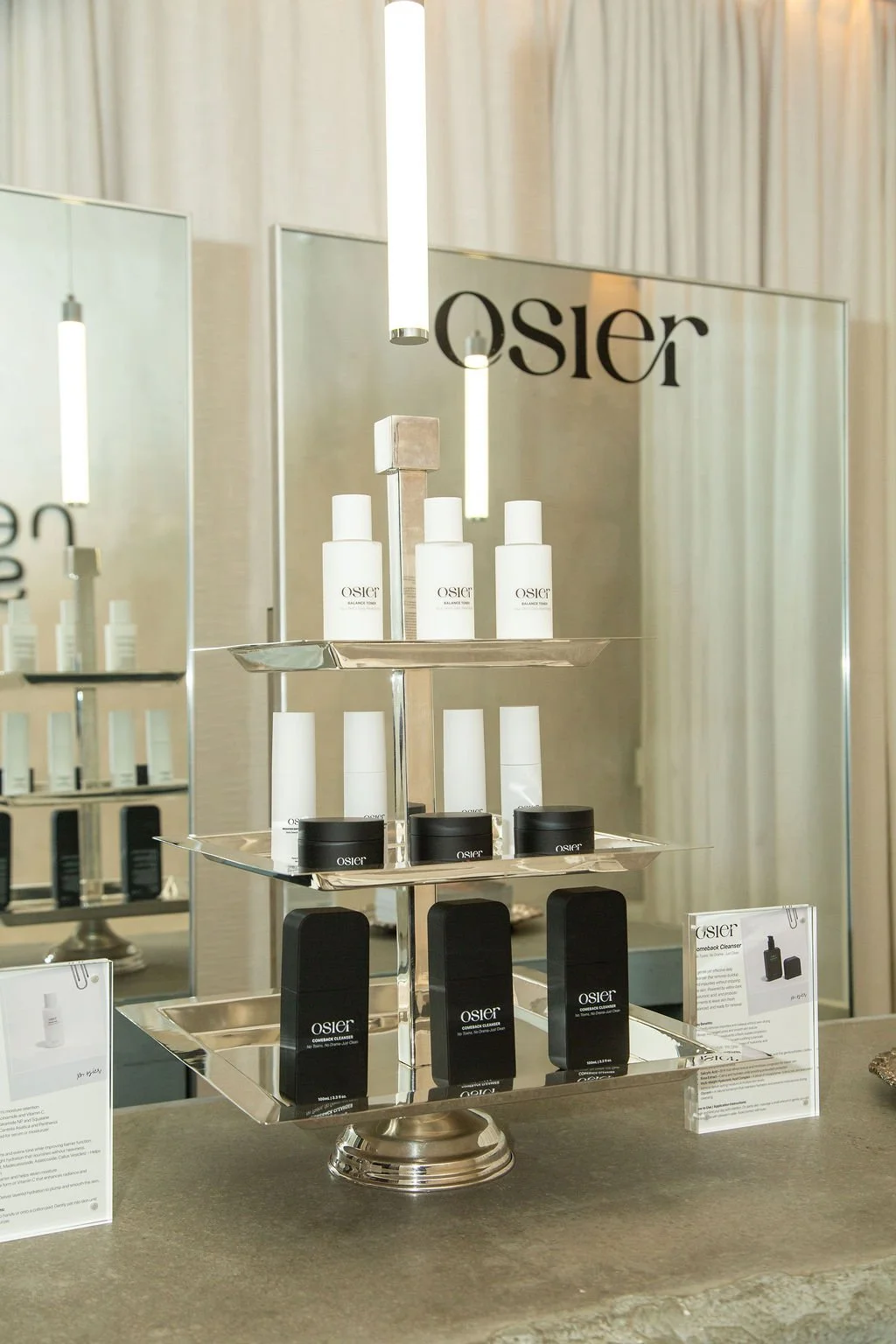 Display of Osier skincare products on a three-tier silver tray, with bottles and boxes arranged neatly, set against a mirror and light-colored curtains.