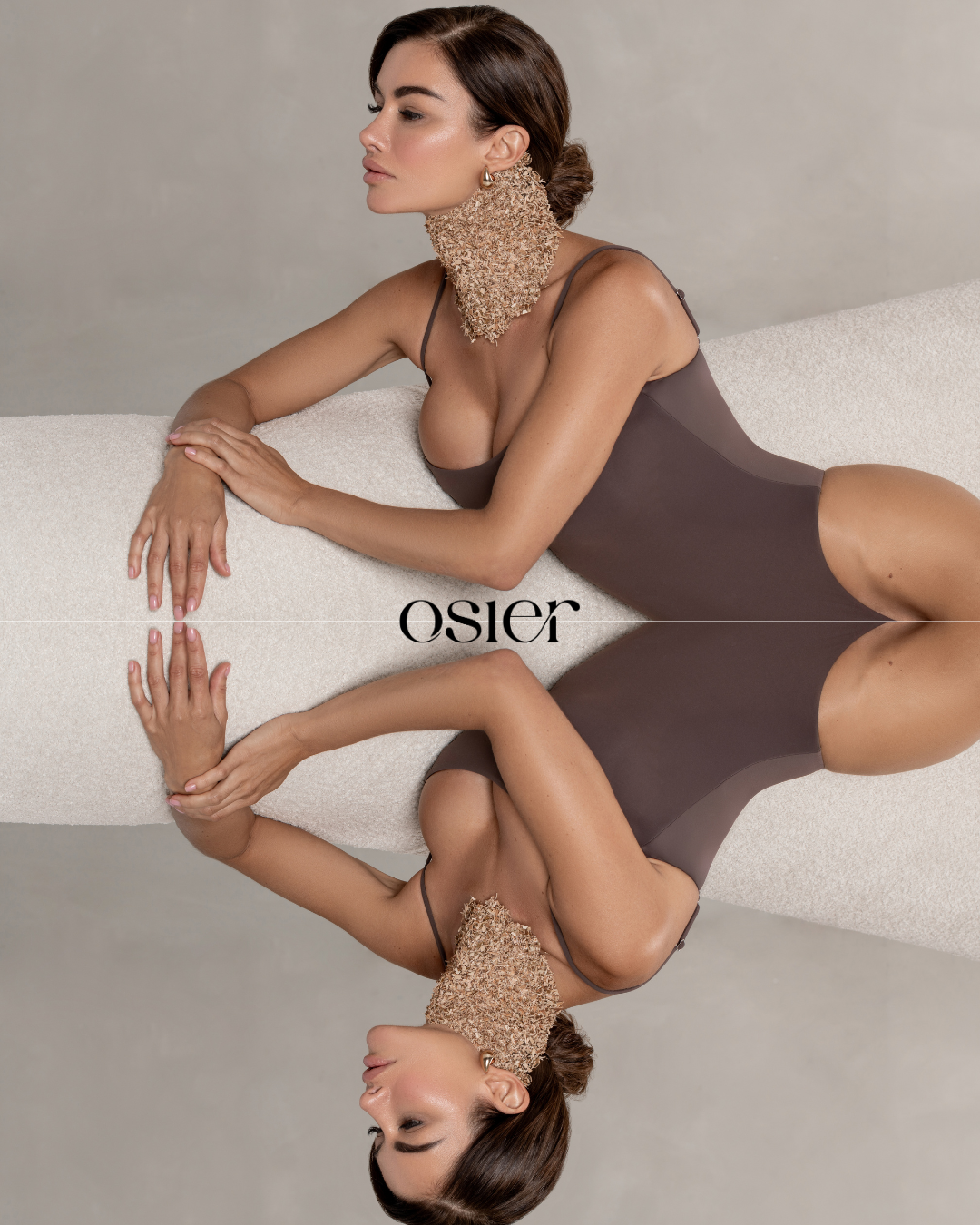 A woman in a brown bodysuit with a textured neckpiece, lying on a beige surface with a mirror reflection, against a plain background.