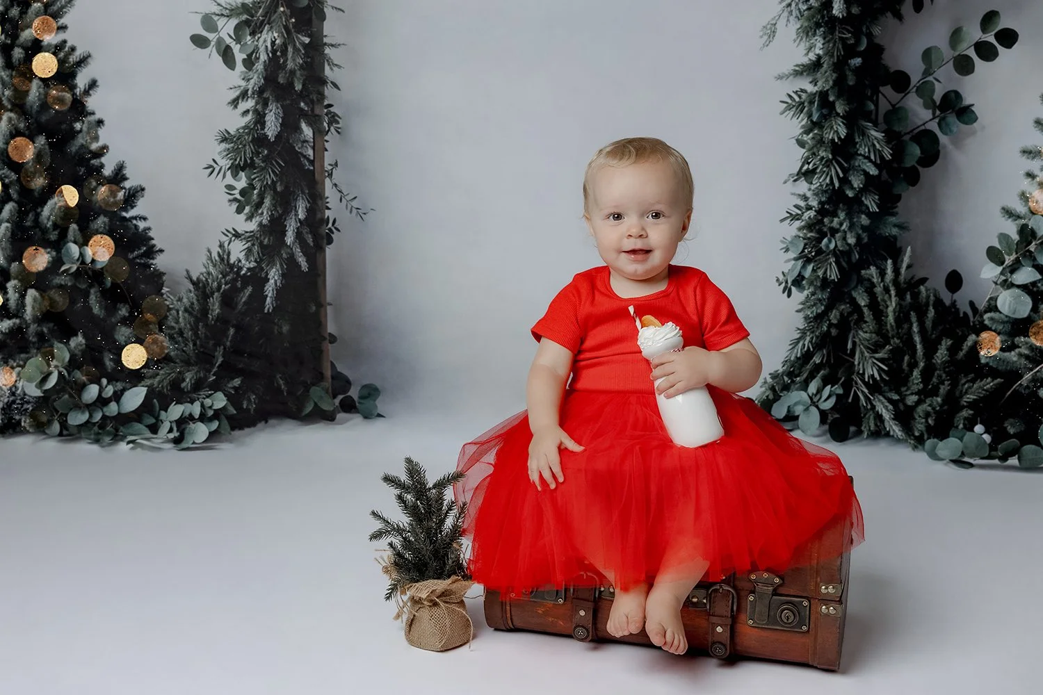 A young girl wearing a red dress sitting on a vintage trunk, holding a milkshake with whipped cream and a straw, with Christmas trees for family christmas photoshoot in the Hill's district in Sydney
