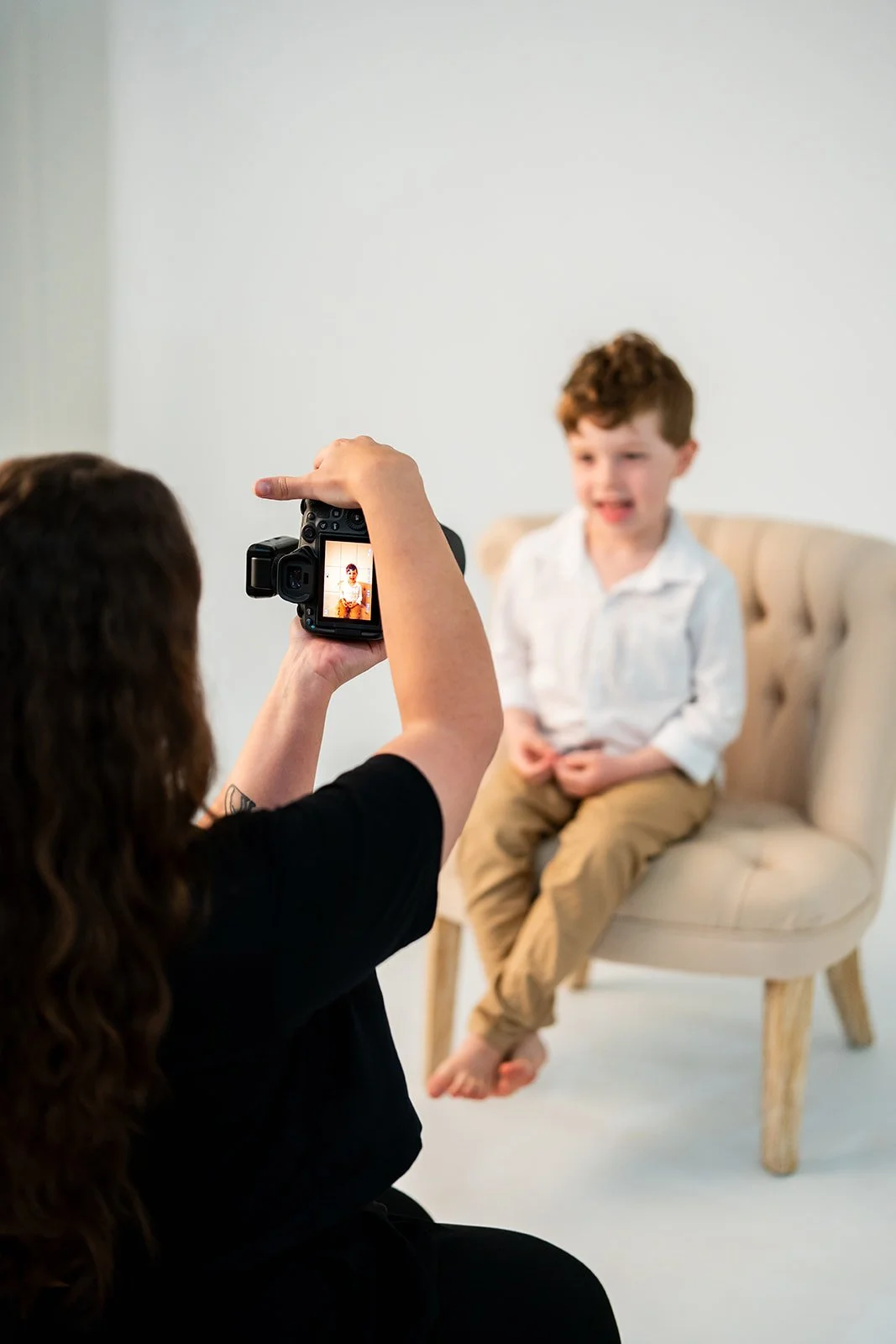 A woman taking a photo of a young boy sitting on a beige chair with a tufted back in a photography studio.