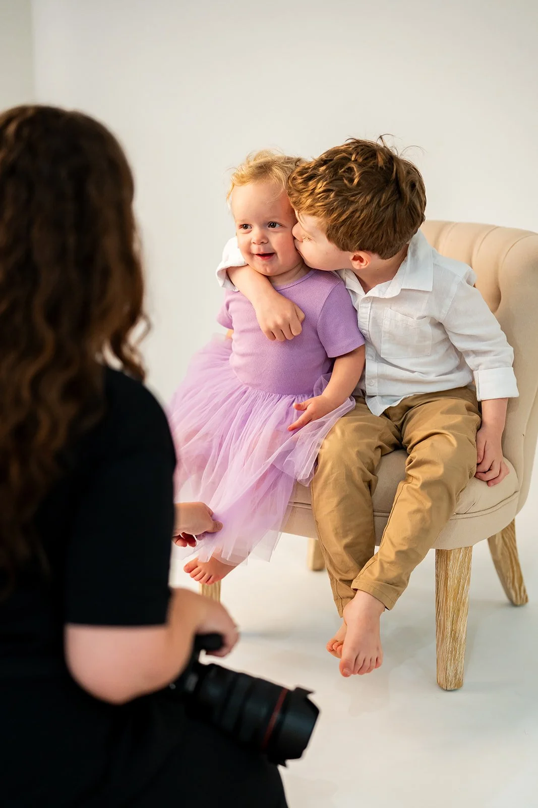 A young boy and girl sitting on a beige armchair, with the boy kissing the girl on the cheek. A woman, partially visible, is holding a camera and facing them, taking their picture.