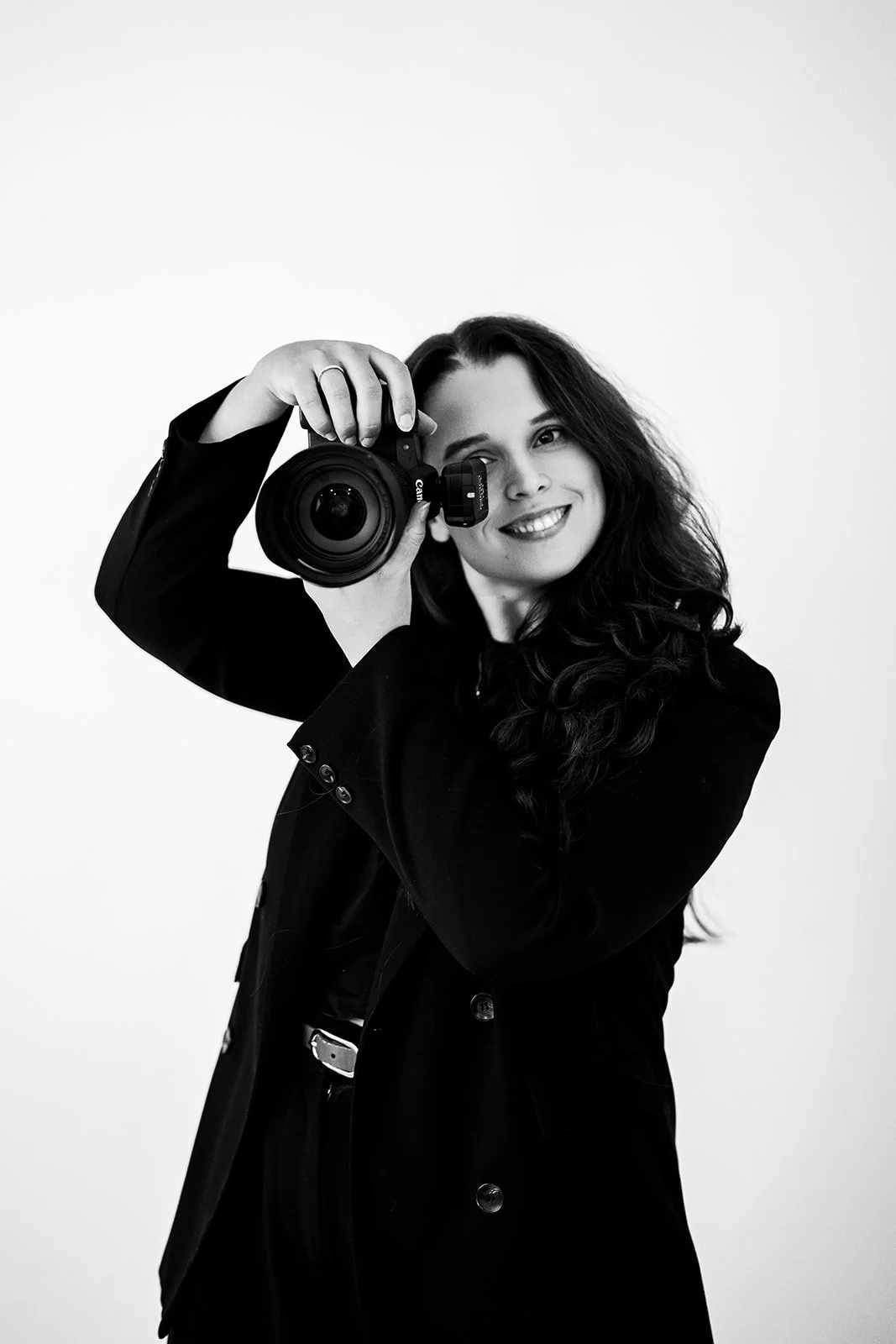 A woman with long curly hair taking a photo with a Canon camera, smiling at the camera, black and white image.