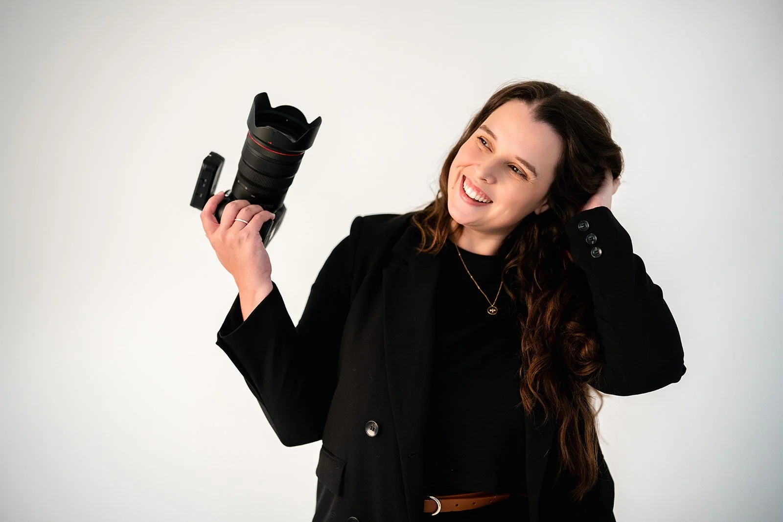 A woman with long brown hair and a black blazer smiling while holding a professional camera with a large lens.