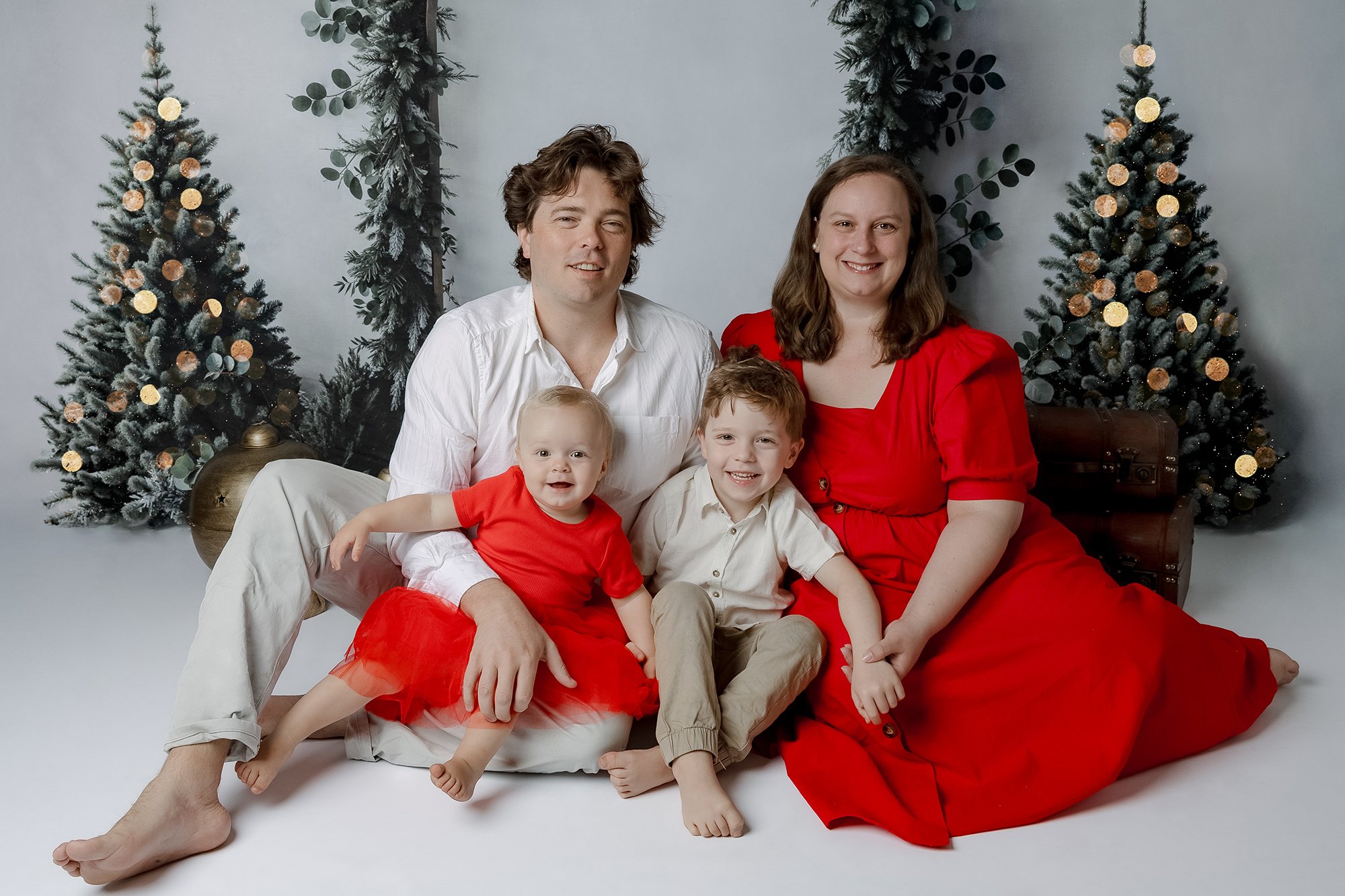 Family of four sitting on the floor with Christmas trees in the background. The father and son are wearing light-colored clothing, while the mother and daughter are dressed in red for a Christmas Family Portrait in Sydney's Hill's District studio.
