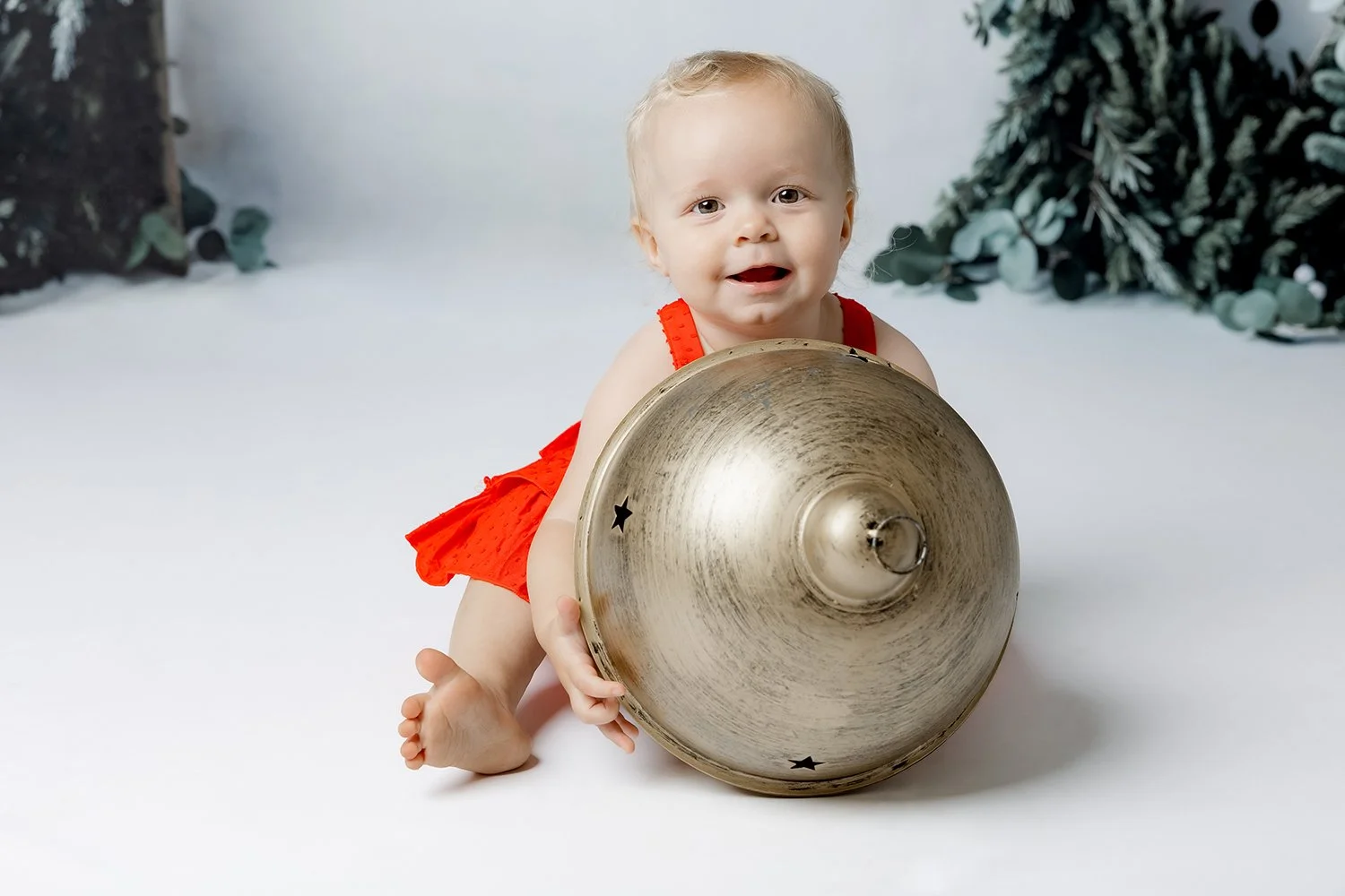 A baby in a red outfit playing with a large vintage metal bell on a white surface with Christmas greenery in the background for a family christmas portrait photoshoot in Castle Hill, Sydney.