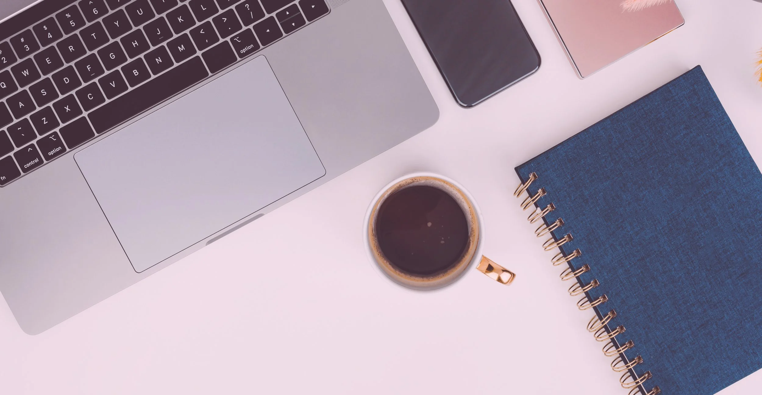Top view of a workspace with a laptop, pen, smartphone, cup of coffee, blue notebook, and some scattered papers on a light pink surface.