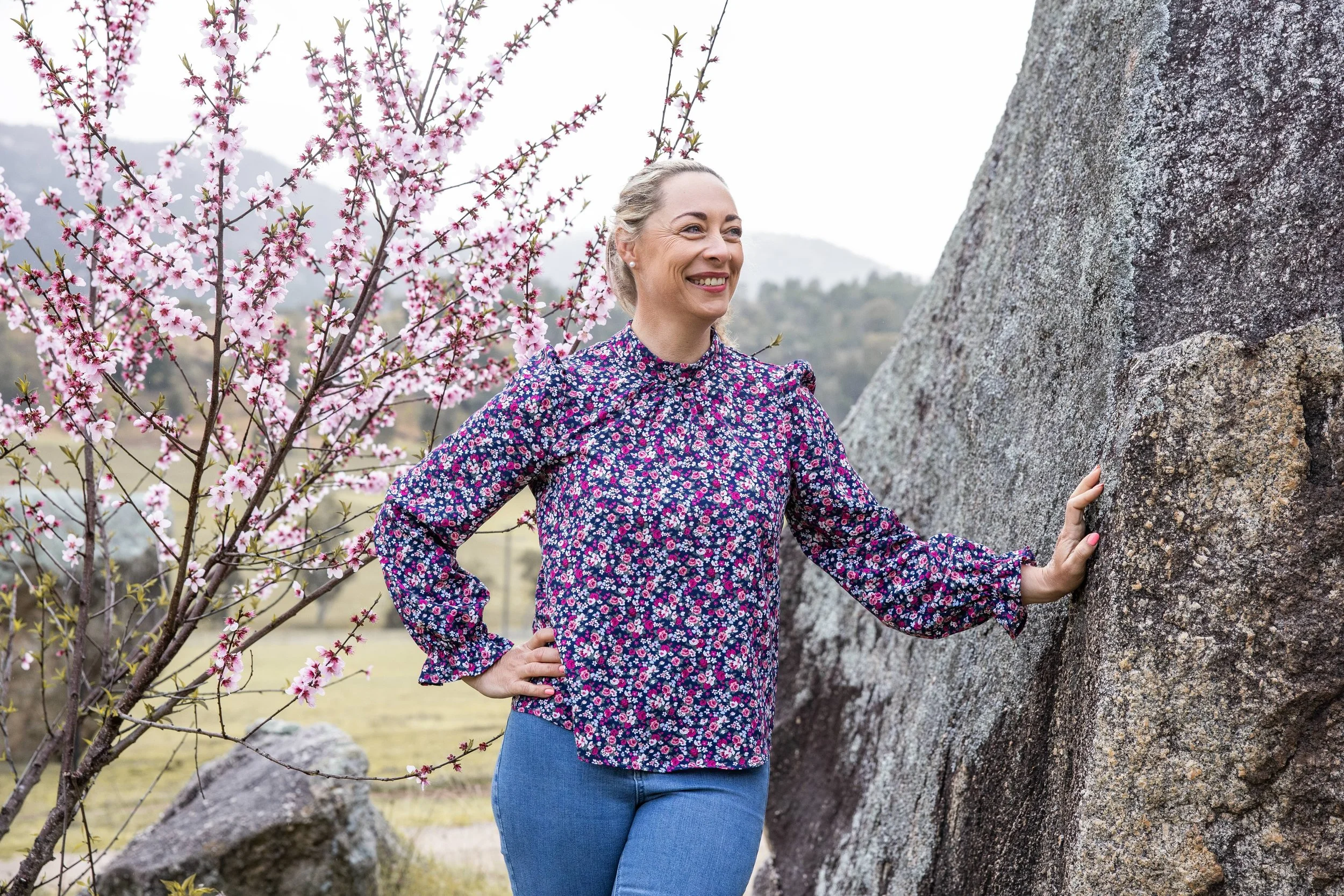 Alex Pope  in a floral blouse standing outdoors near a large rock, smiling, with pink blooming trees and hills in the background in Tamworth, NSW