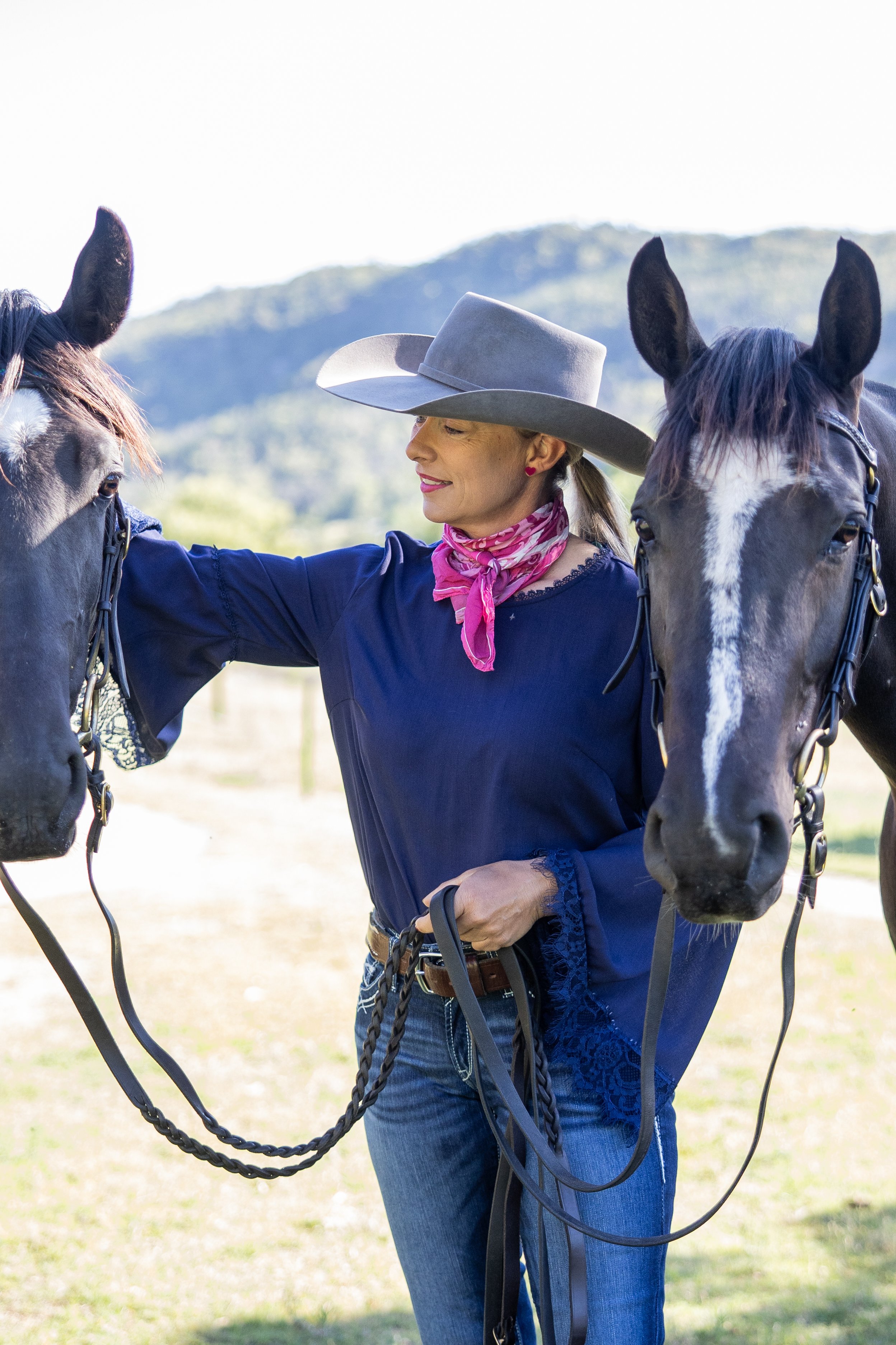 A woman wearing a hat, pink scarf, and blue blouse standing between two horses outdoors with a mountain in the background.