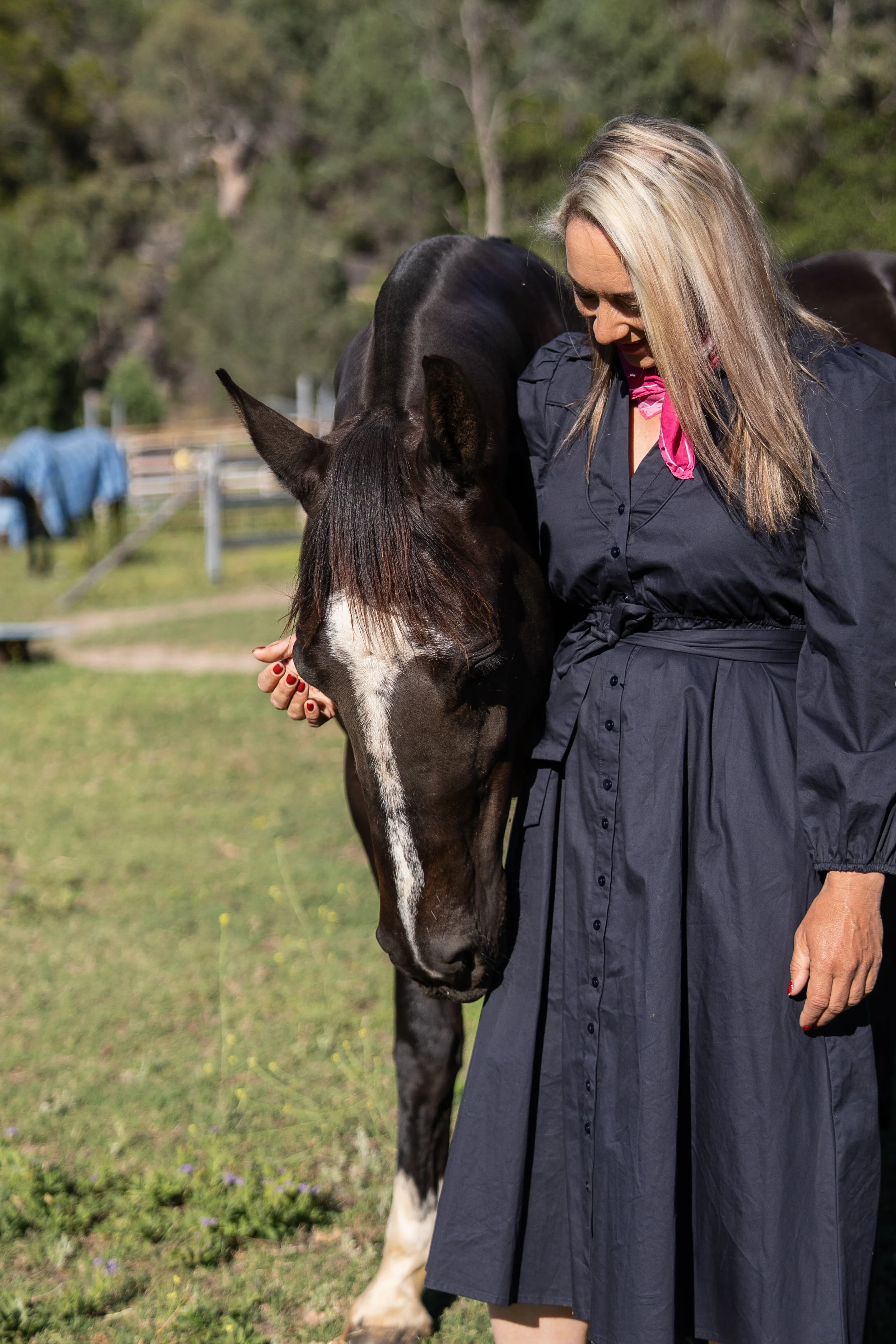 Alex Pope with blonde hair in a navy blue dress and pink scarf gently touching a black horse with a white blaze on its face, standing on a grassy field with trees in the background.