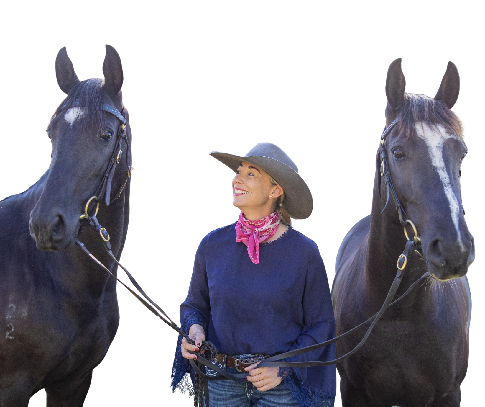 A woman wearing a wide-brimmed hat and a pink scarf standing between two black horses with bridles, holding their reins and smiling.