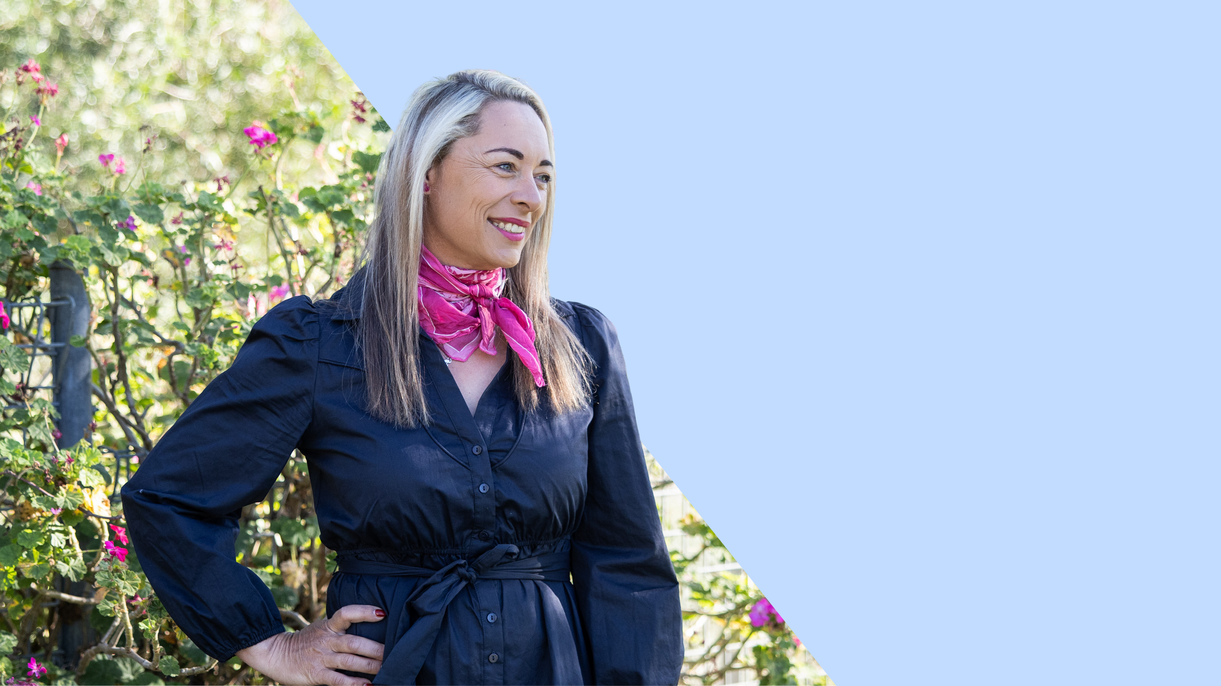 Alex Pope workplace HR consultant with blonde hair, wearing a navy blue dress with a pink scarf, standing outdoors in front of flowering bushes.