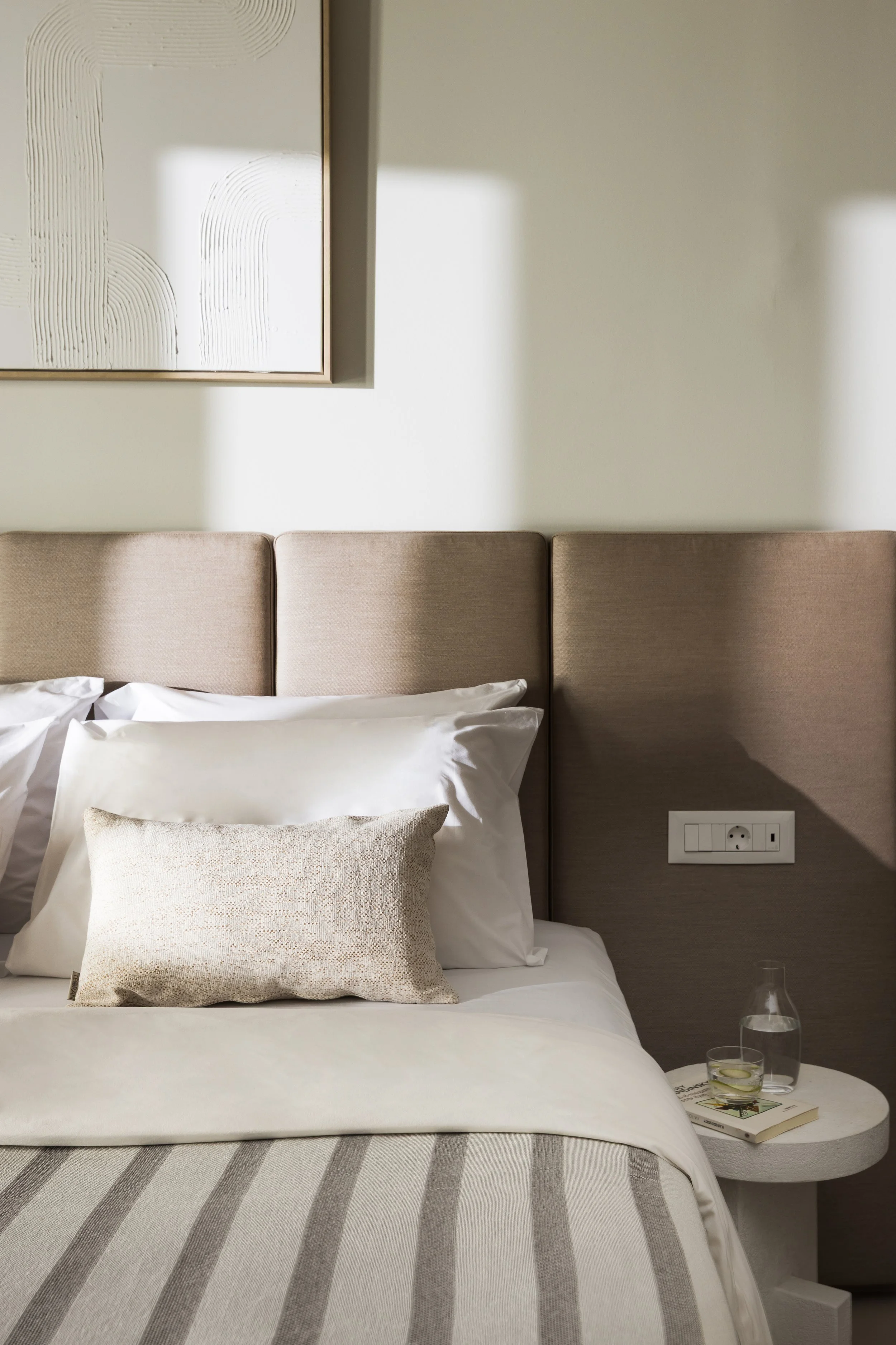 A neatly made bed with white and beige pillows, a striped bedspread, and a beige padded headboard. A white side table holds a glass of water, a pitcher, a book, and a small plate with a slice of lemon. Sunlight casts shadows on the wall and headboard.