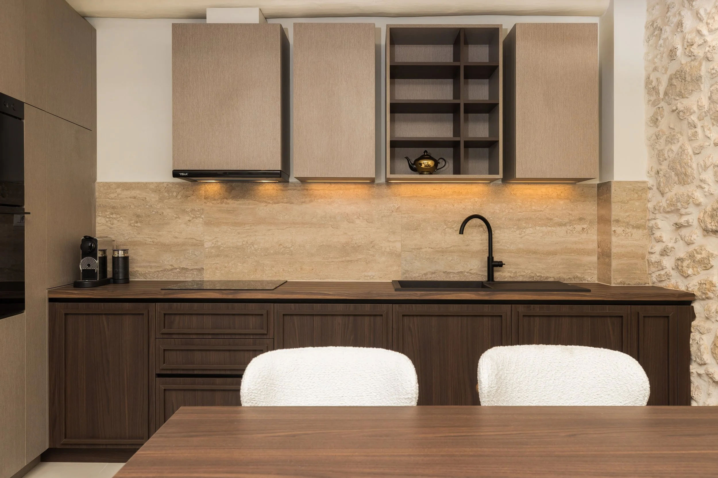 Modern kitchen with dark wood cabinets, beige stone backsplash, open shelves, black faucet, coffee machine, teapot, and white upholstered chairs in the foreground.