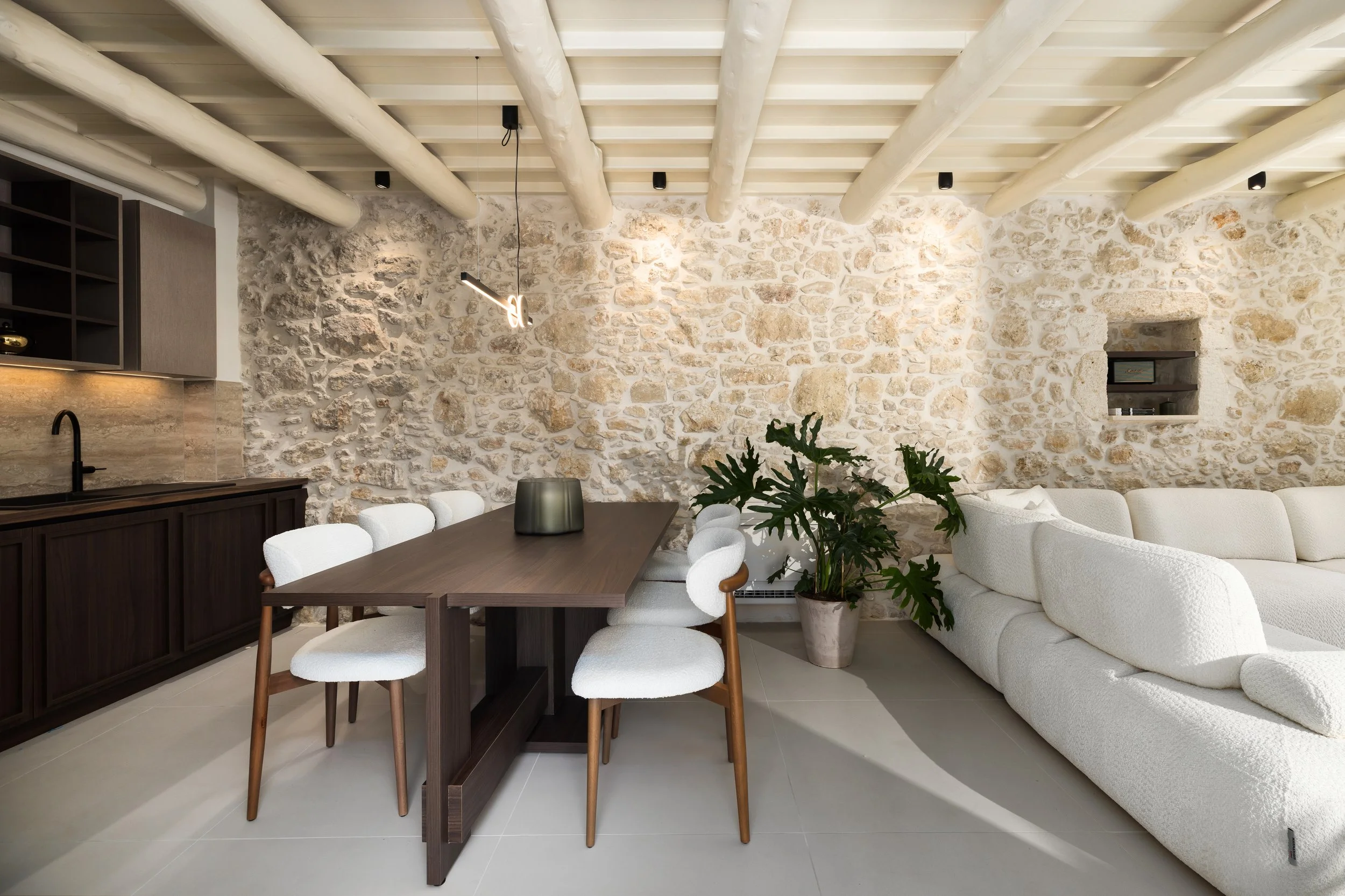 Modern living room with stone wall, white sofa, wooden dining table with white upholstered chairs, potted plant, kitchen area with black sink, and ceiling beams.