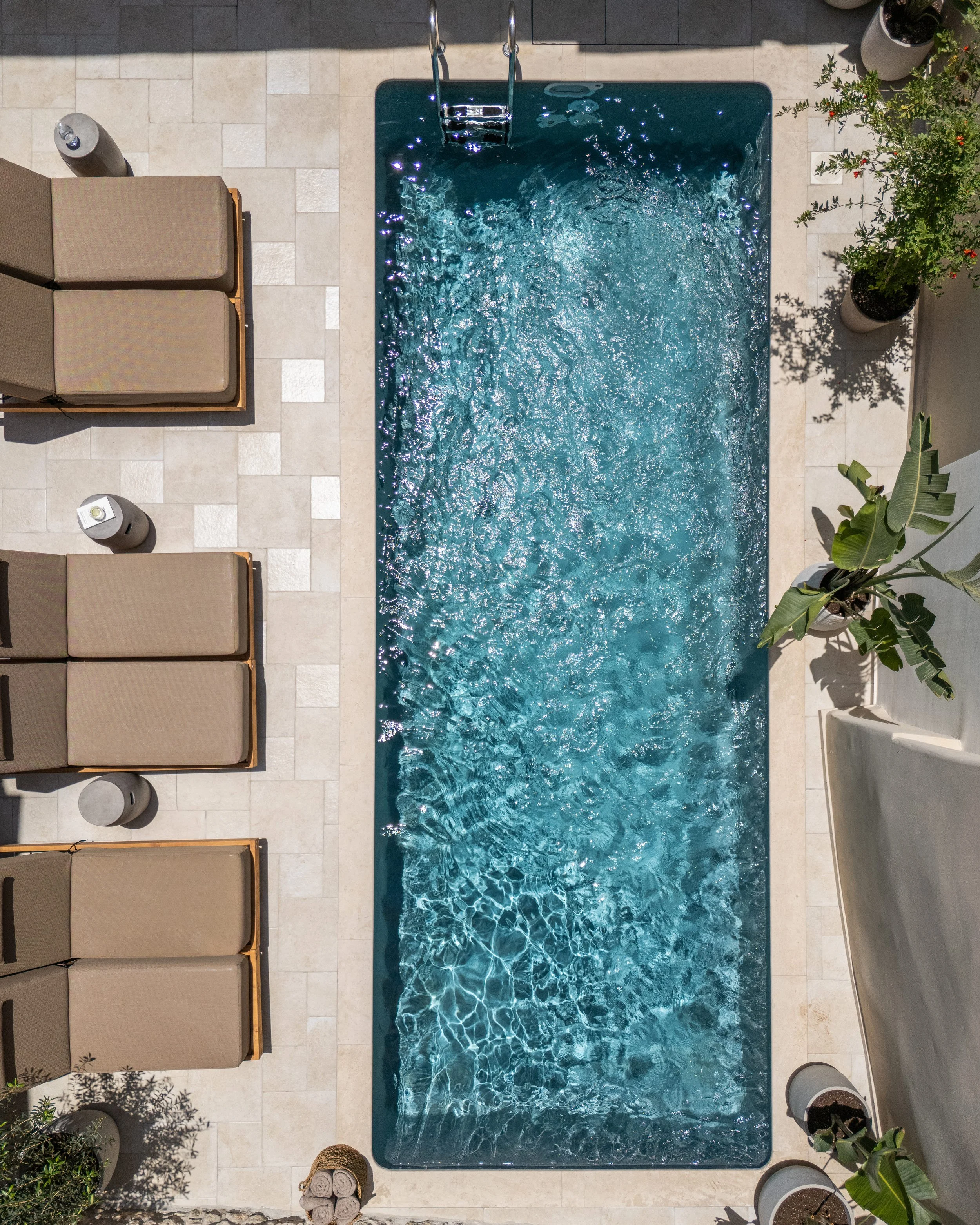 An aerial view of a rectangular swimming pool with light-colored surrounding tiles, lounge chairs on the left, potted plants on the right, and a ladder at the top of the pool.