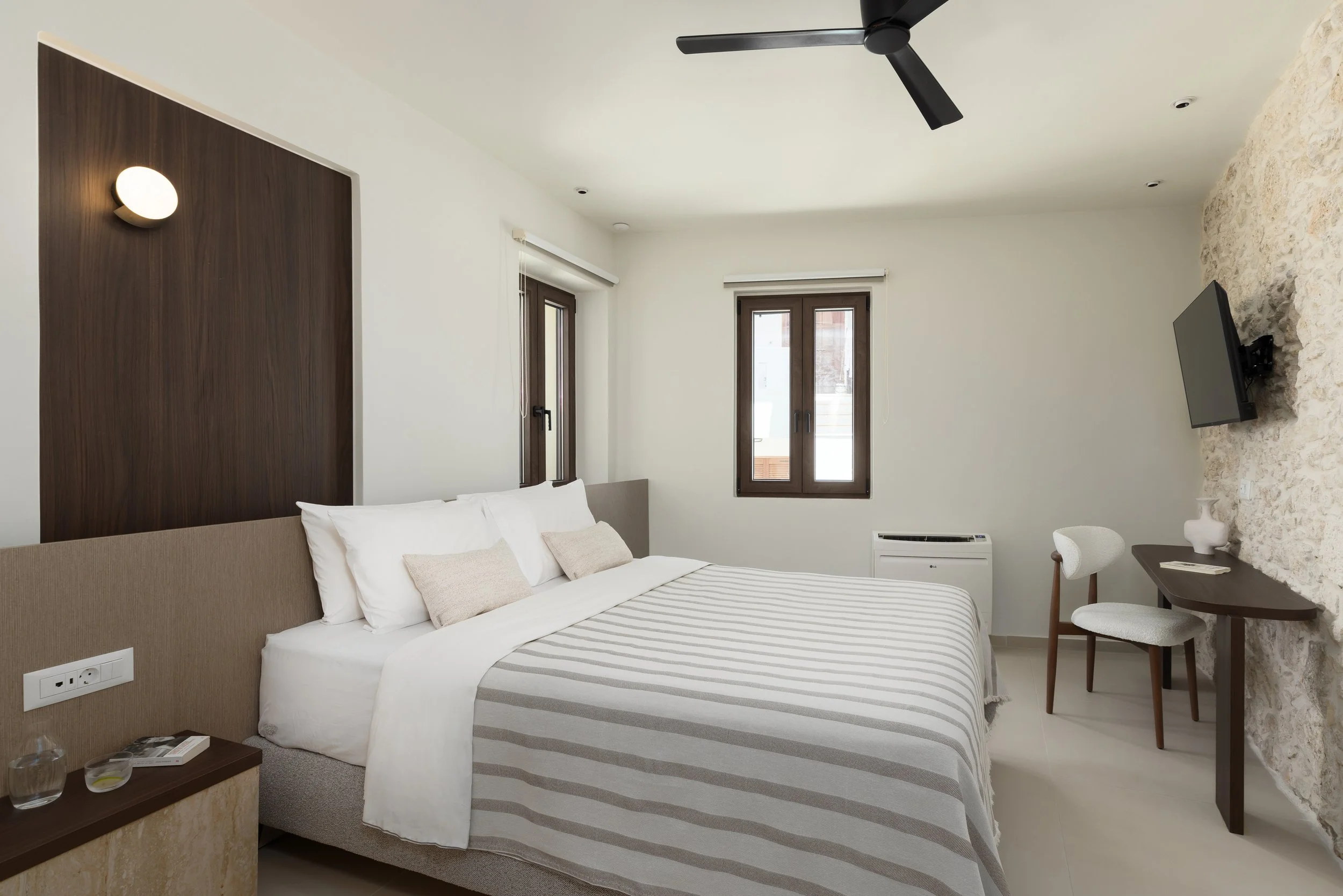 A modern hotel room with a bed, beige headboard, white bedding, and beige pillows. There are two windows with dark brown frames, a wall-mounted TV on a textured stone wall, a small dark wooden desk with a white chair, and a ceiling fan. The room has minimal decor and neutral colors.