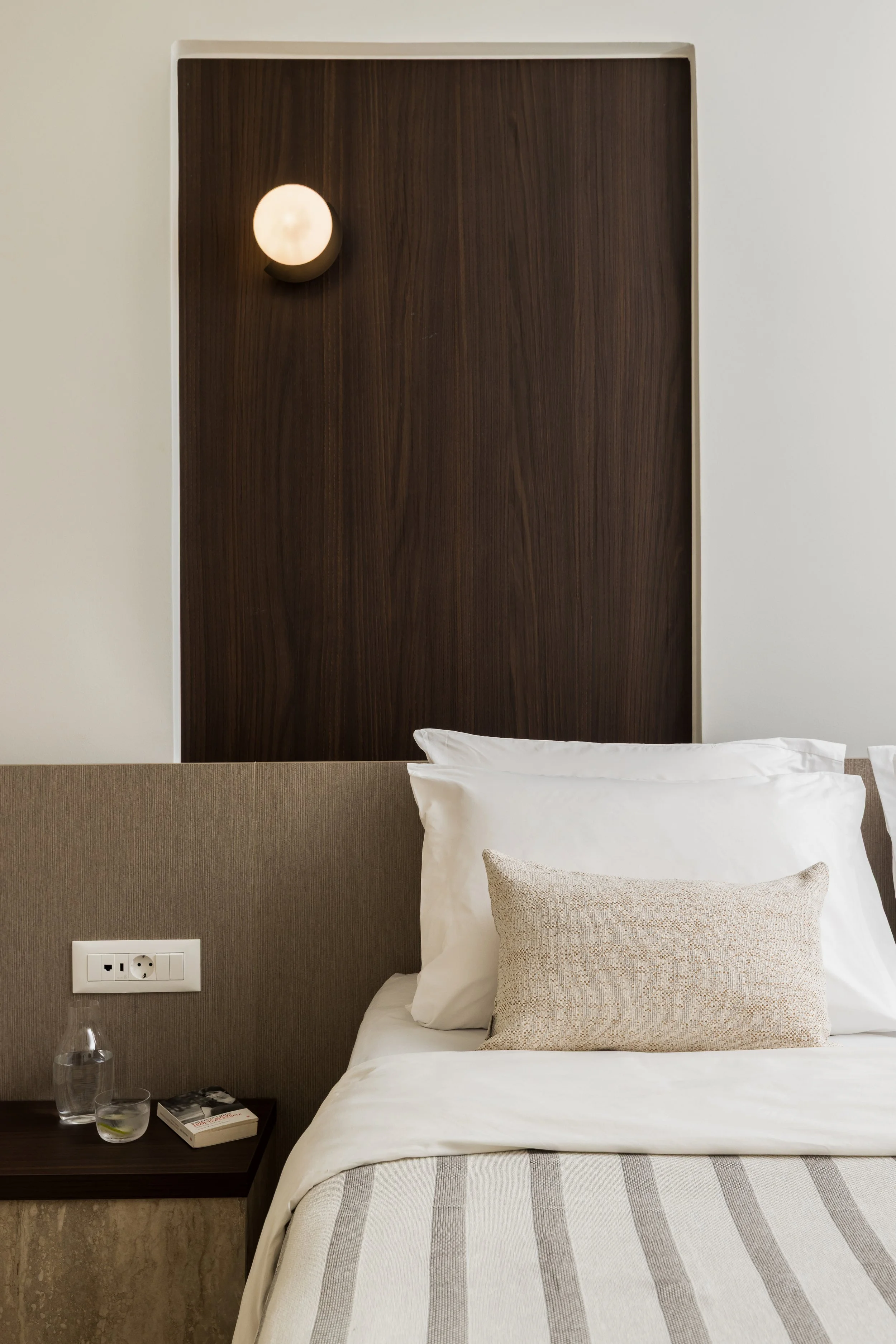 A hotel room with a bed featuring white pillows and a beige pillow, a bedside table holding a glass of water, a book, and a water pitcher, a wall socket, and a lit wall lamp on a dark wooden headboard against a light wall.