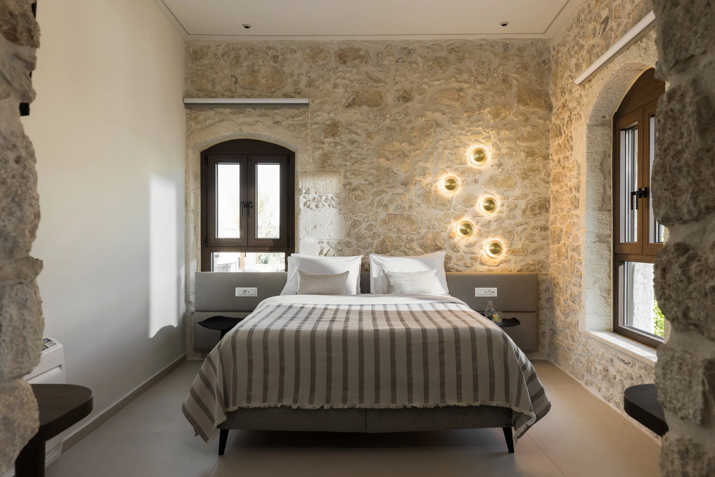 Bedroom with stone walls, bed with white and beige bedding, bedside tables, and unique wall-mounted light fixtures.