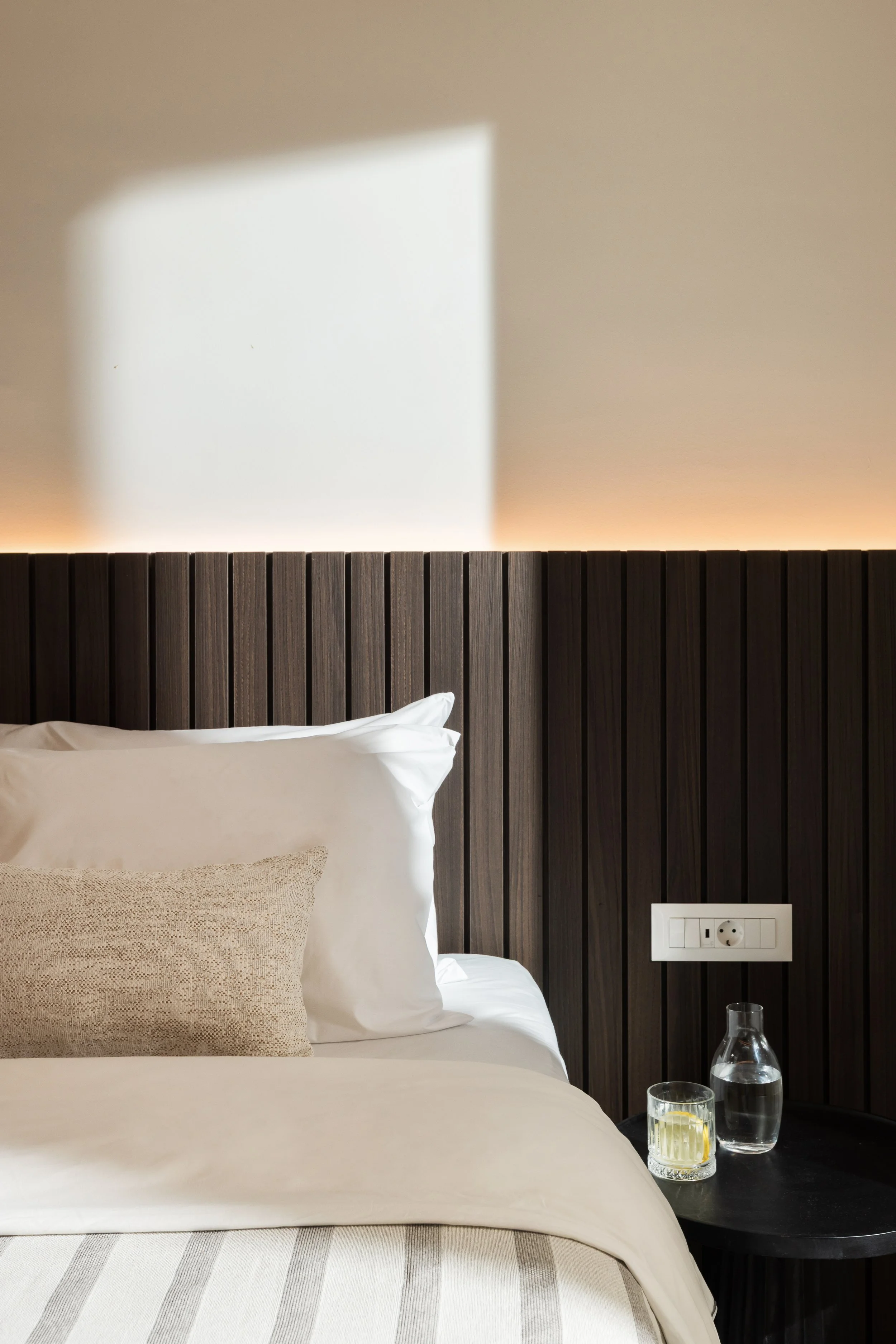 Close-up of a neatly made bed with white pillows and beige accent pillow, dark wooden headboard, and a side table with a glass of water, a glass of lemon water, and a water pitcher in a cozy, modern bedroom.