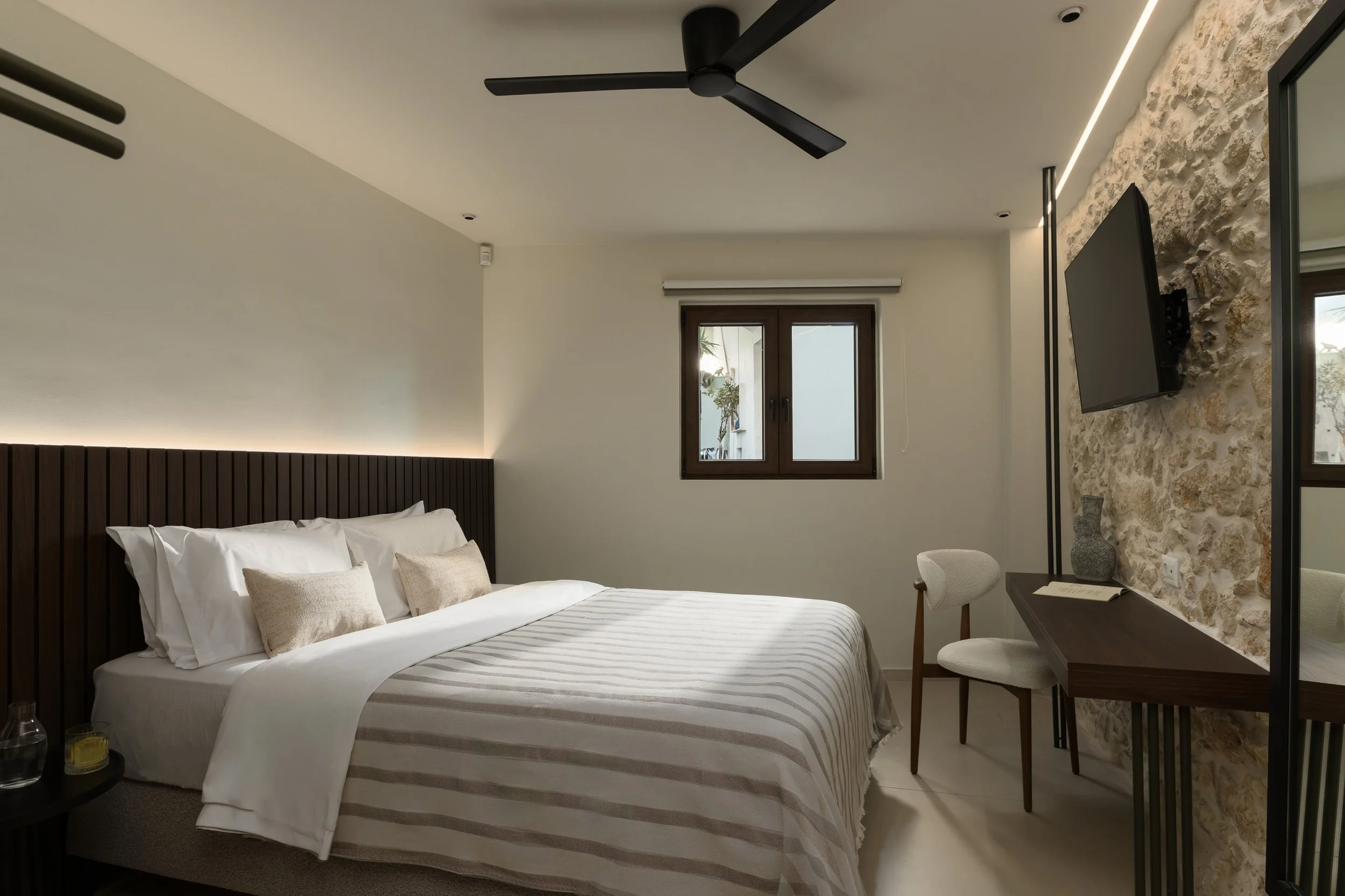 Modern bedroom with a large bed, white bedding, beige pillows, dark wooden headboard, a window, and a stone accent wall with a mounted flat-screen TV.