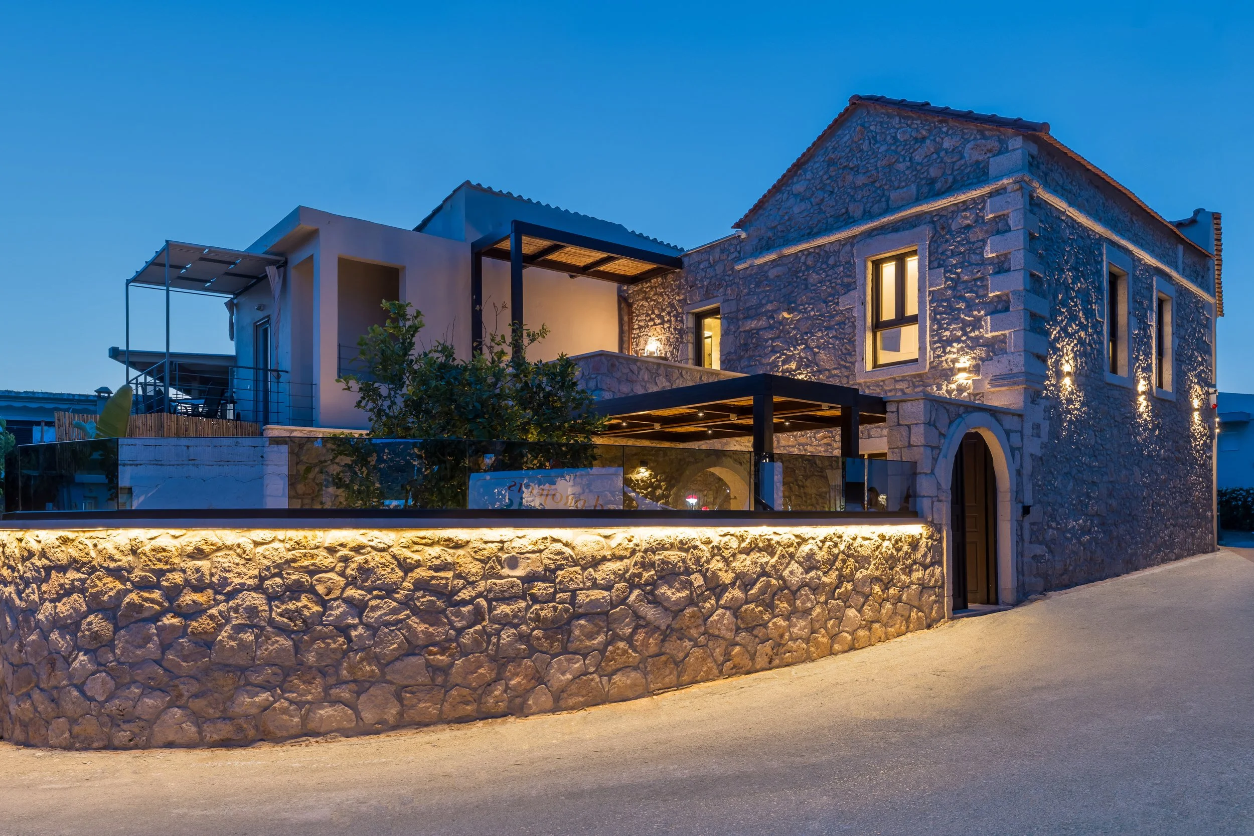A modern stone house at dusk with exterior lighting, featuring arched doorways, multiple windows, a balcony, and outdoor seating area, surrounded by a decorative stone wall.