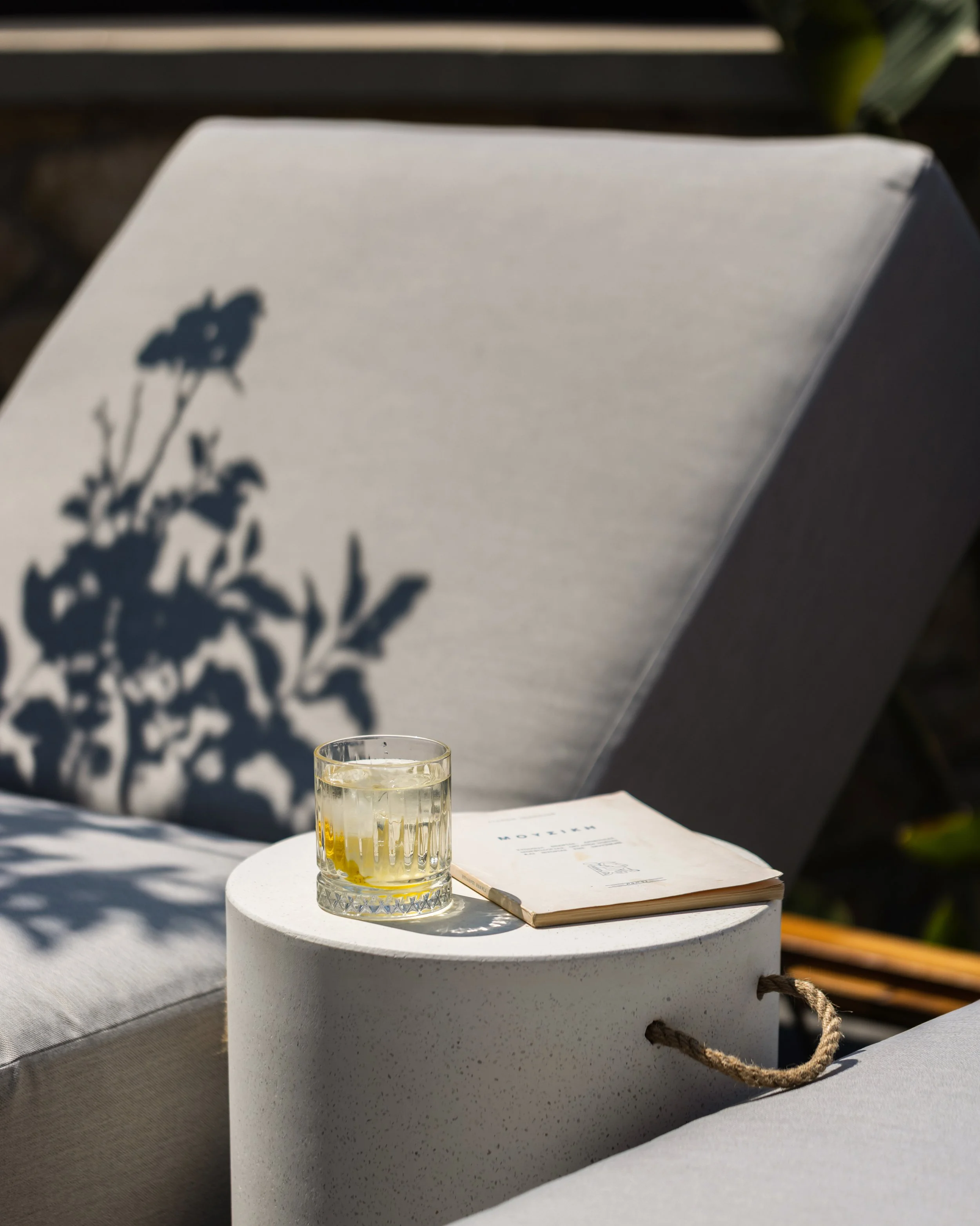 A glass of iced tea and a notebook sit on a small white table outside, with soft shadows of a plant cast on a lounge chair in the background.
