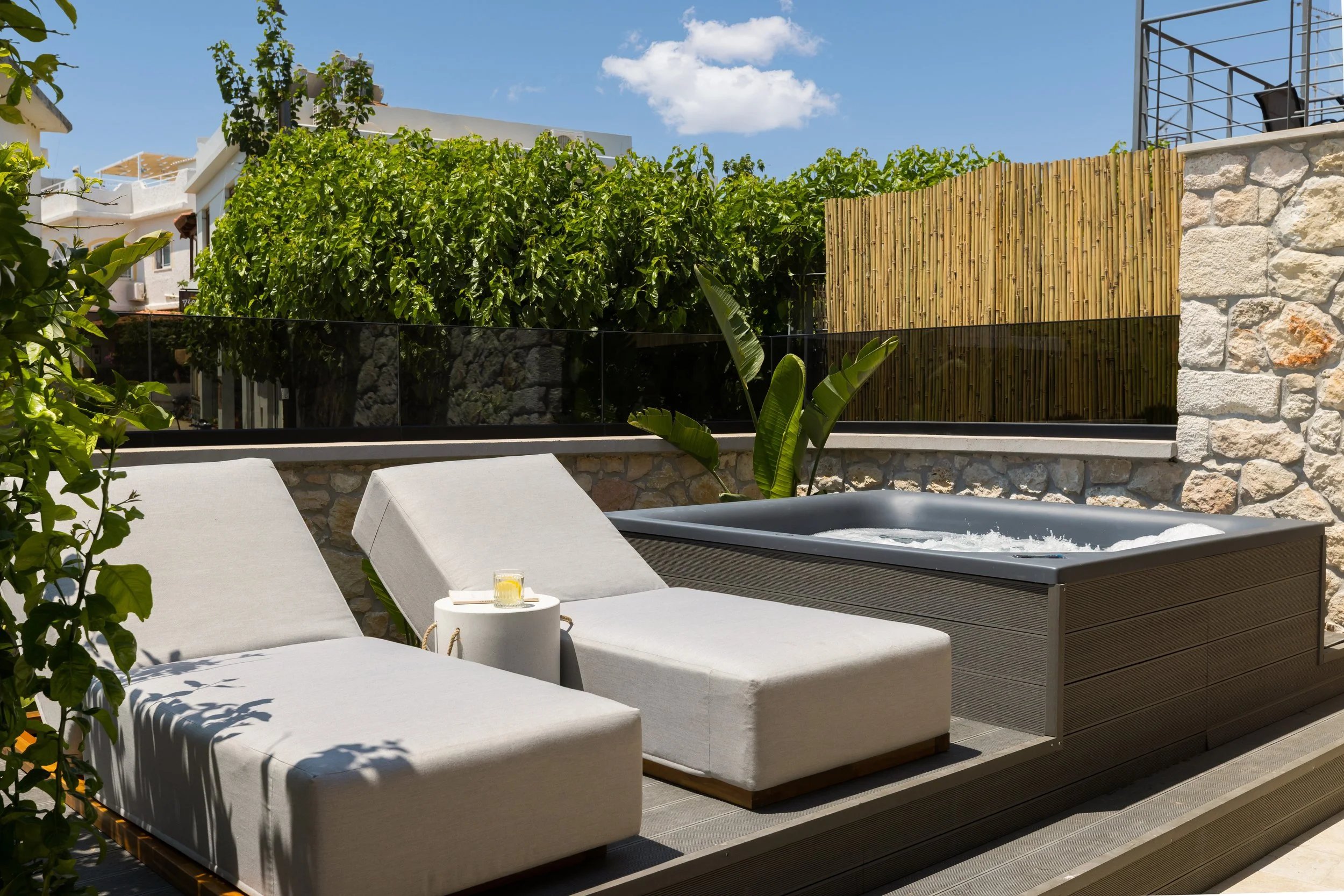 Outdoor patio with two white lounge chairs, a small round table, a hot tub, lush green plants, and a stone wall with a bamboo fence in the background.
