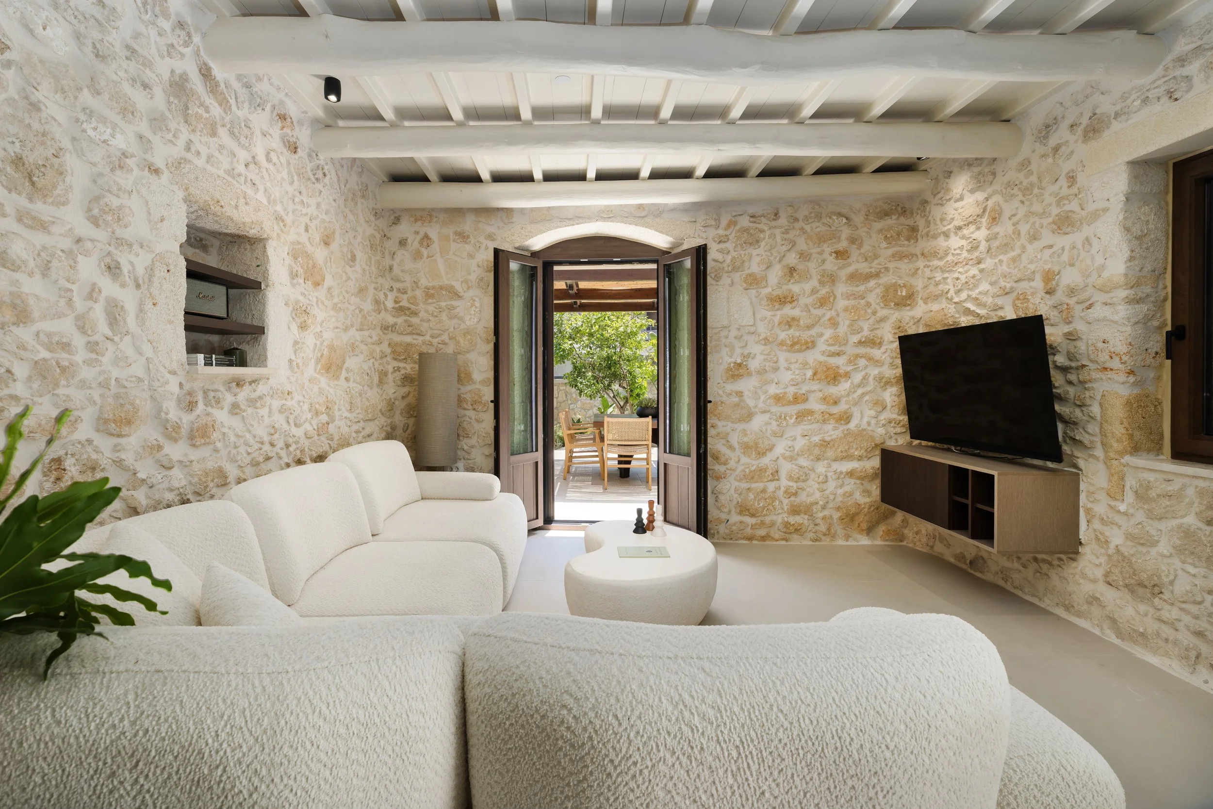Living room with stone walls, white textured sofas, a round white coffee table, a mounted flat-screen TV, and open double doors leading to a patio with outdoor furniture and greenery.