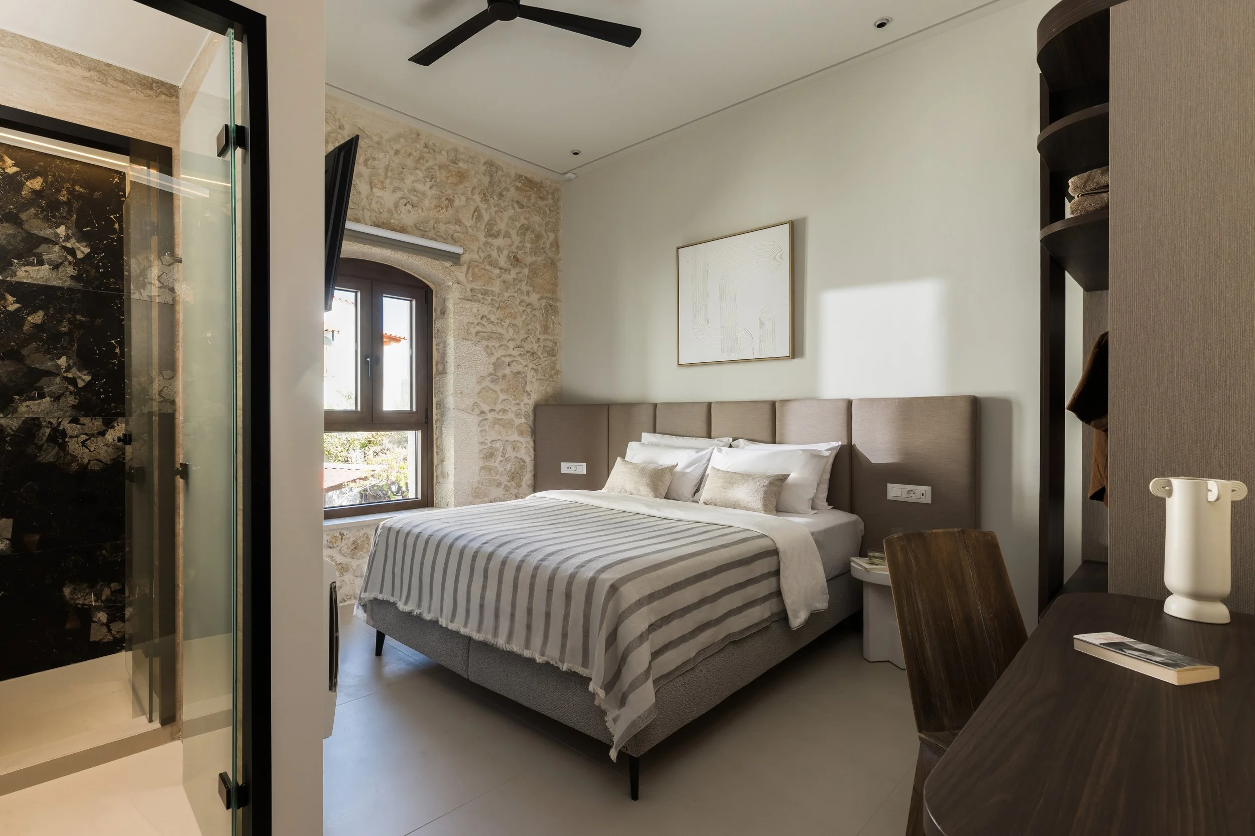 Modern hotel bedroom with stone and painted wall, window, bed with beige headboard, white bedding, and striped blanket, wooden desk with a book and white vase, open closet with clothes, ceiling fan, and television.