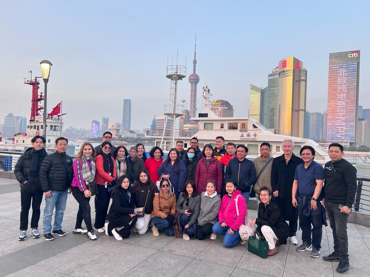 Group of 21 people smiling and posing for a photo on a waterfront promenade, with a large boat and Shanghai skyline featuring the Oriental Pearl Tower and other skyscrapers in the background.