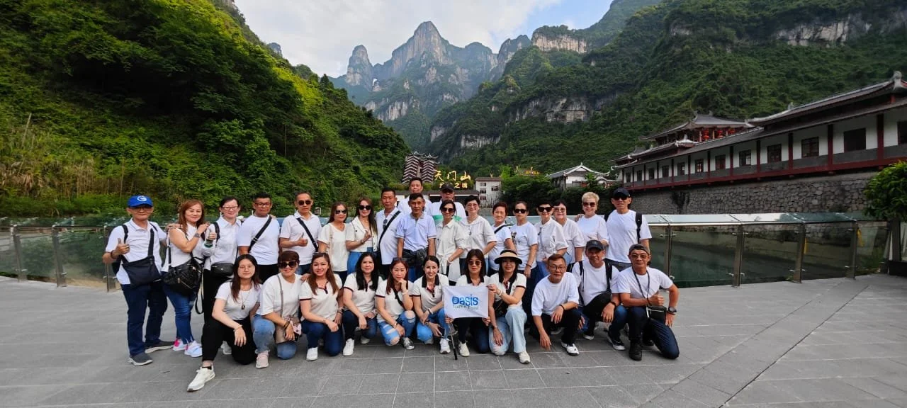 Group of people in white shirts posing for a photo outdoors, mountains with lush greenery in the background, traditional building on the right side.