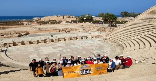 Group of tourists holding a large orange banner inside an ancient outdoor amphitheater with a view of the sea in the background.
