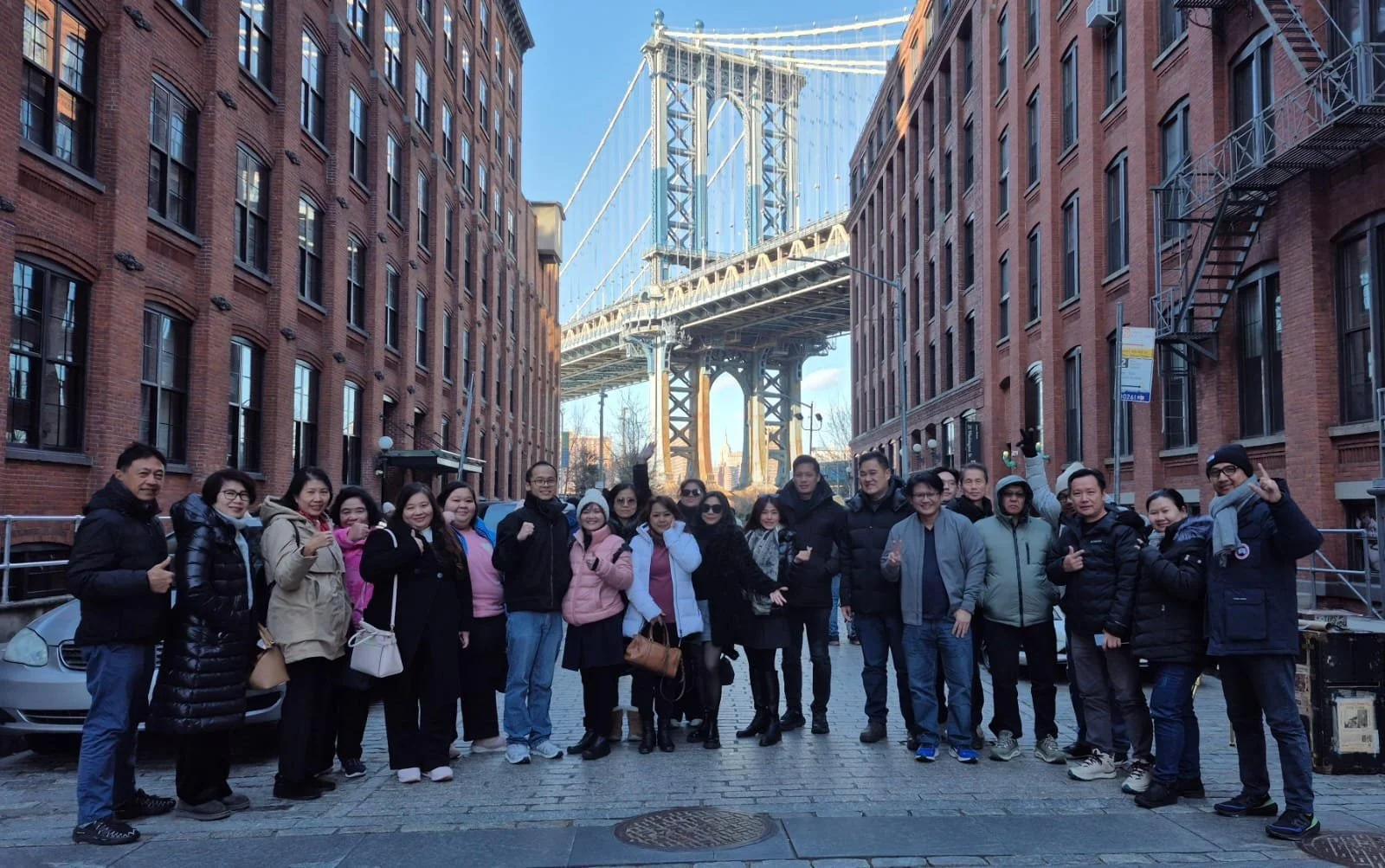 Group of people standing on cobblestone street in front of a bridge, with red brick buildings on both sides and clear blue sky above.