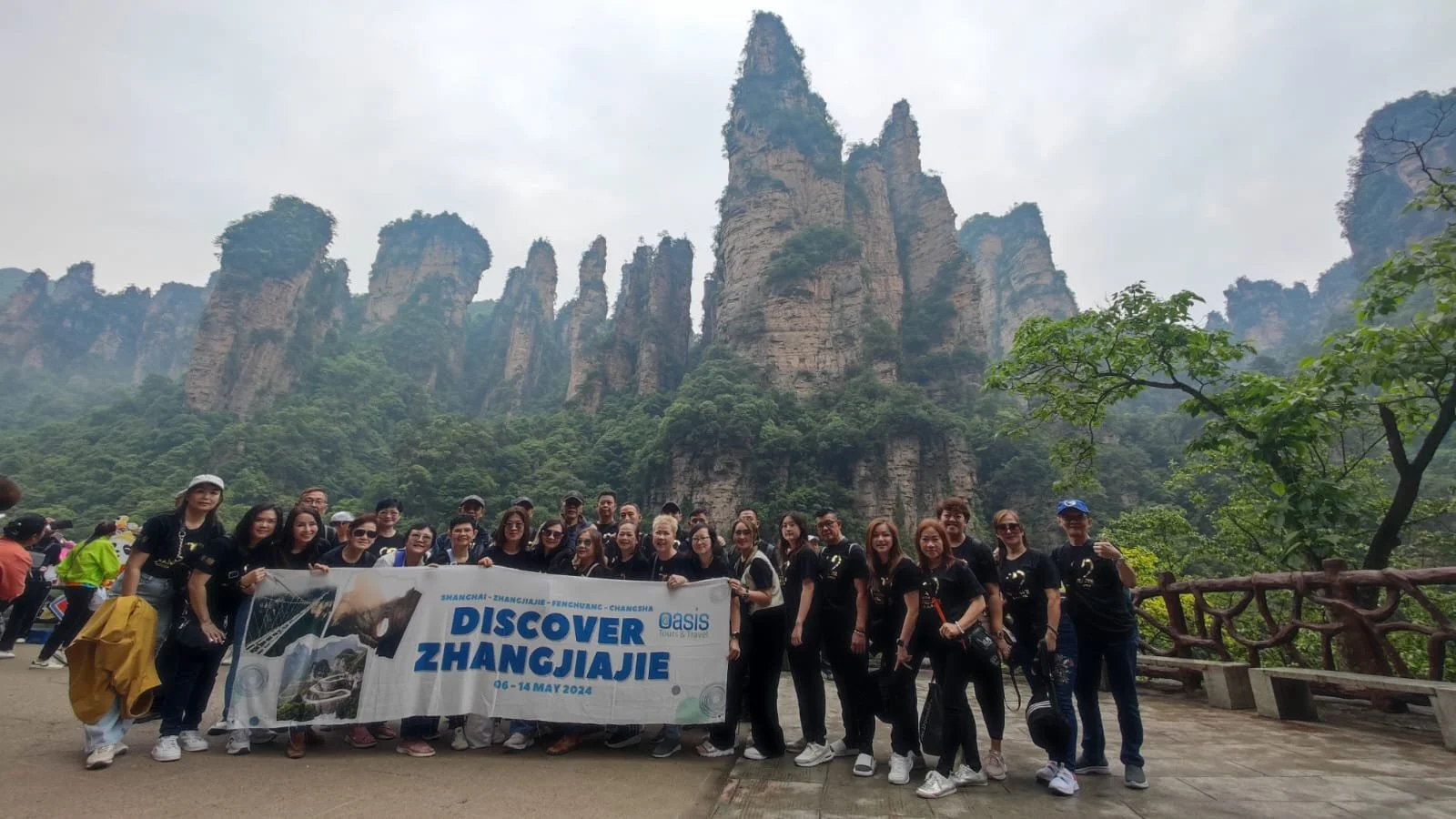 Group of tourists holding a banner that says Discover Zhangjiajie, standing in front of tall, rocky mountain formations covered in greenery.