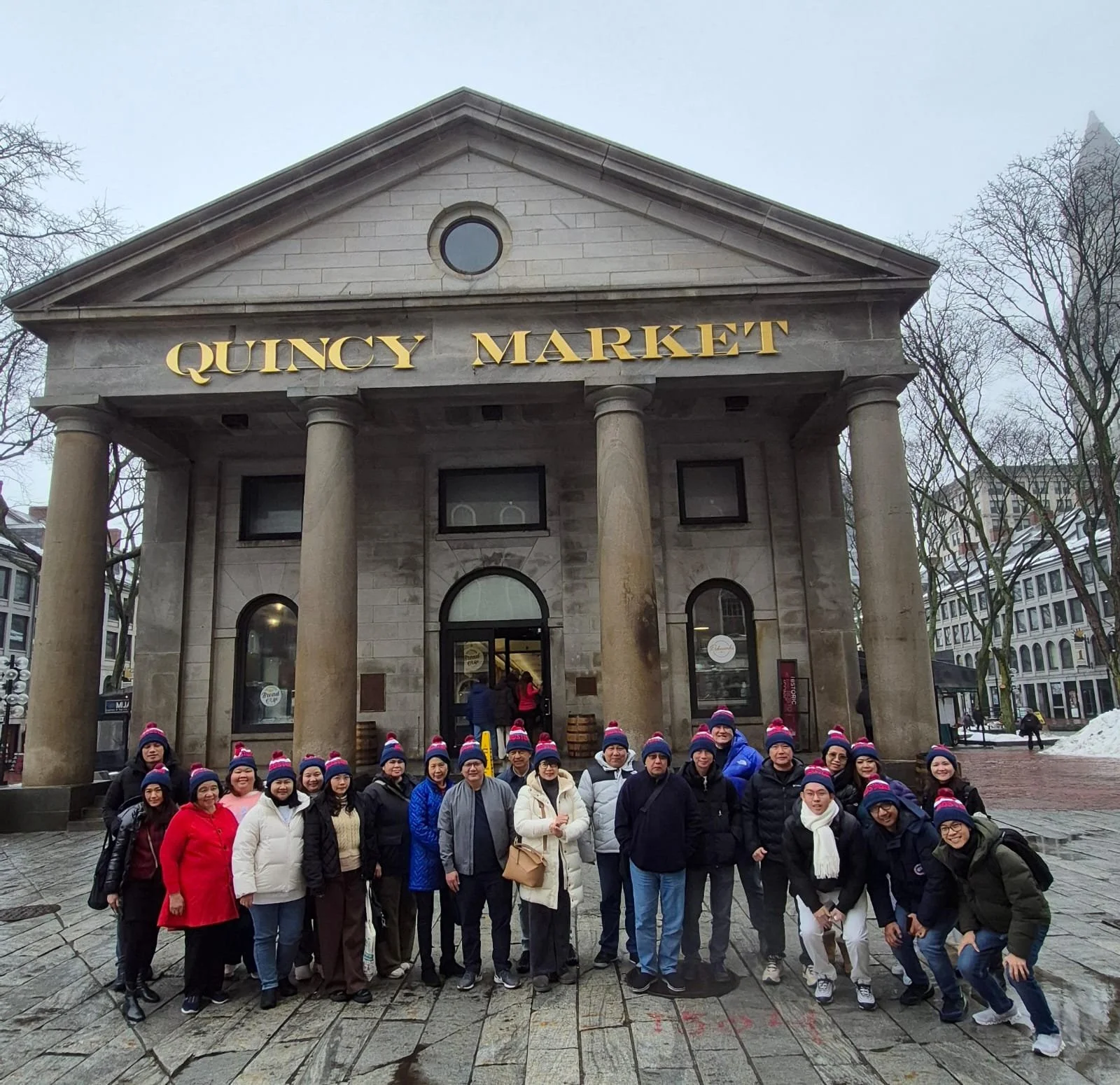 Group of people wearing pink, purple, and white knit hats standing in front of Quincy Market building with tall columns, stone facade, and gold lettering.
