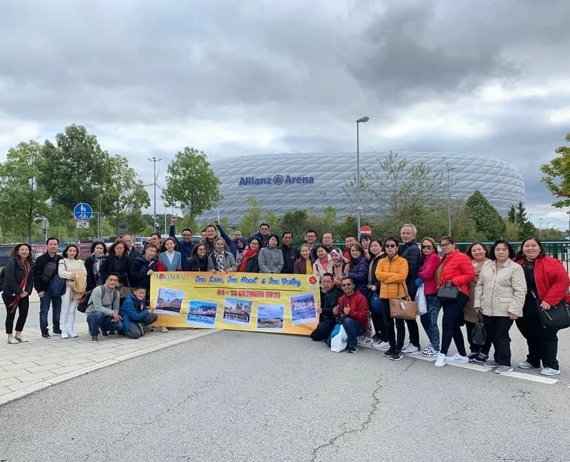 Group of people standing outside Allianz Arena, holding a yellow banner with images of a castle and text, with the stadium in the background.