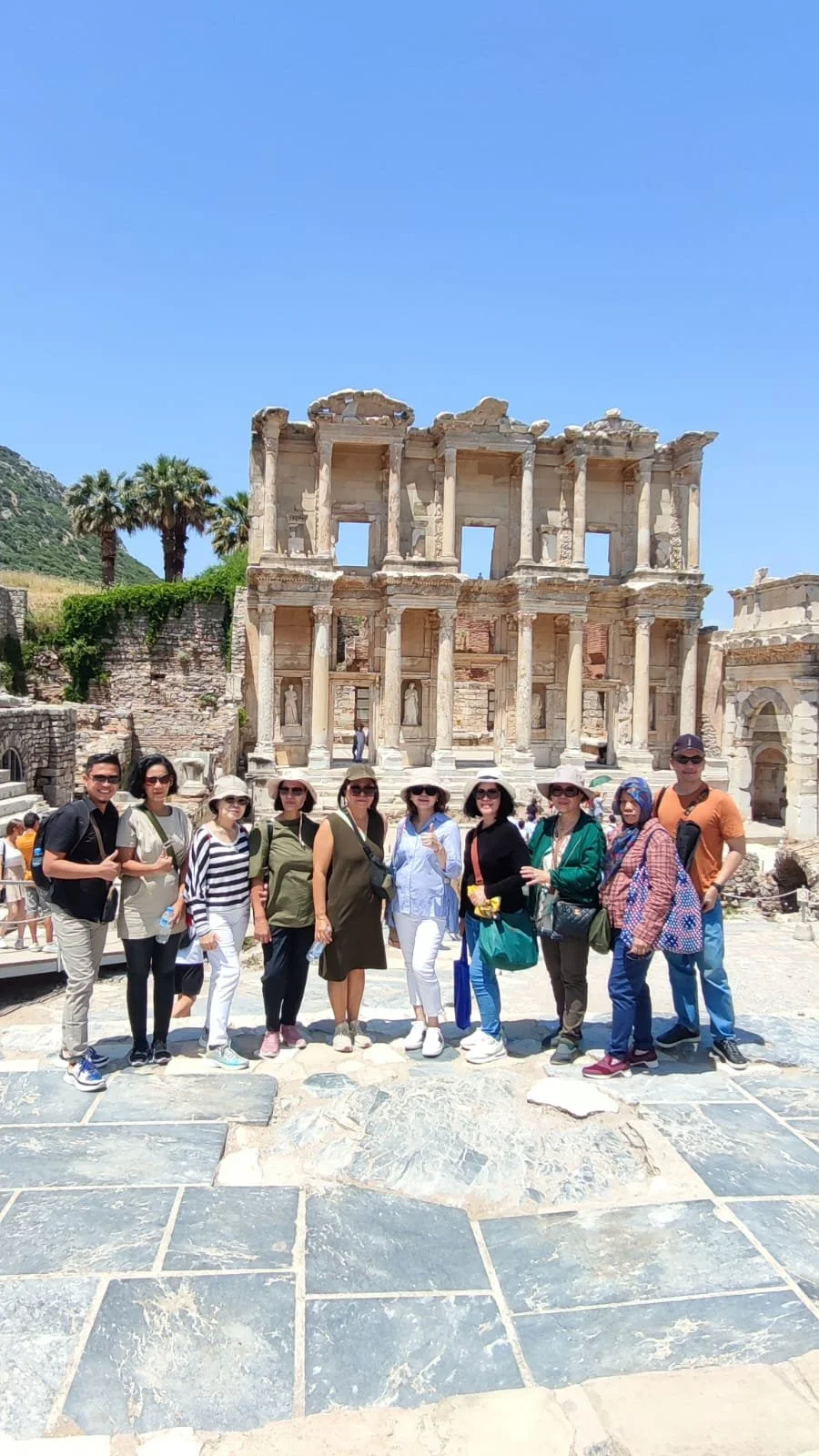 A group of tourists standing in front of an ancient Roman archaeological site with a large, two-story stone building with columns and intricate carvings under a clear blue sky.