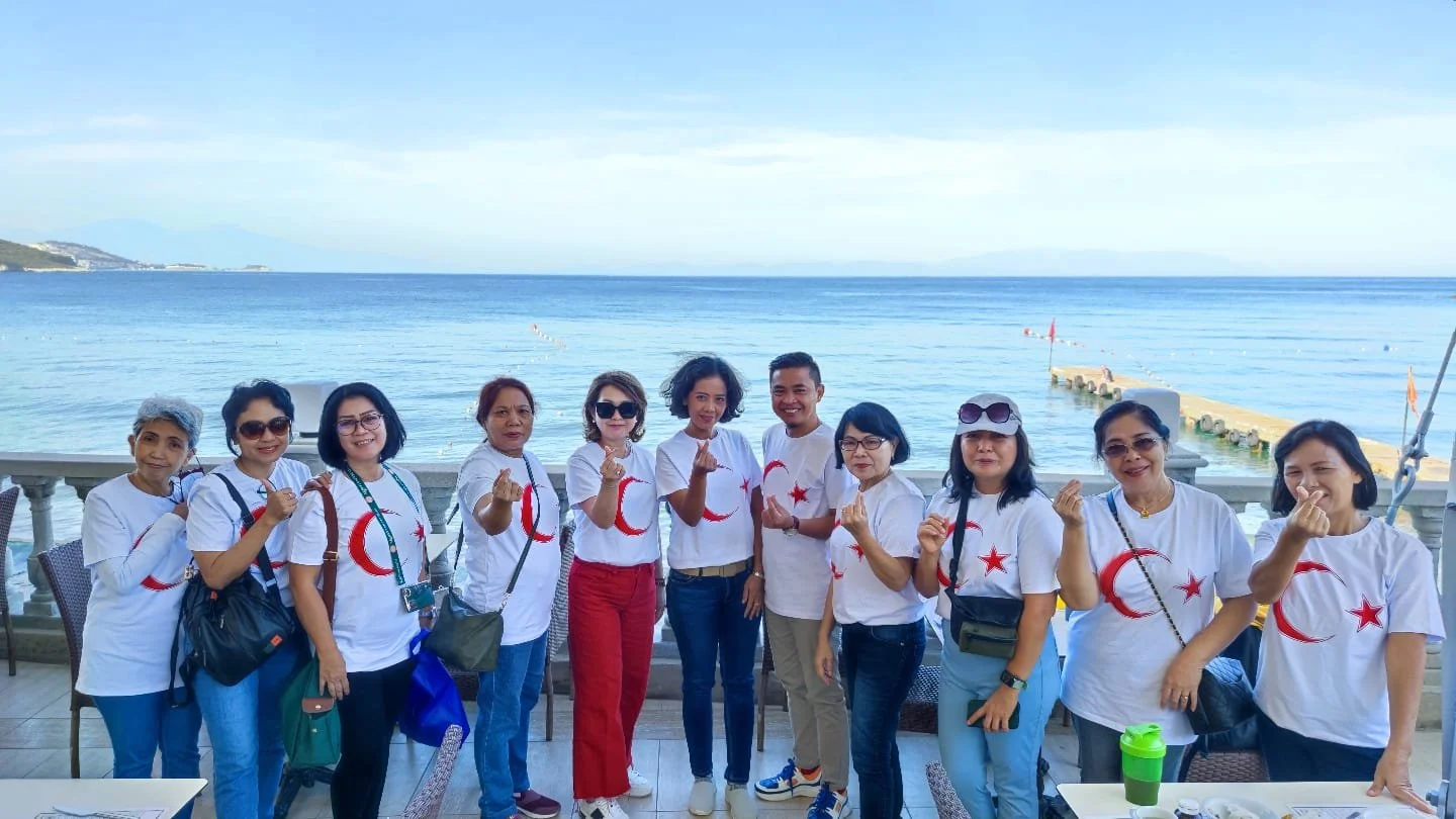 Group of women standing on a restaurant patio by the water, wearing matching white t-shirts with red crescent and star symbols, making finger heart gestures, with a scenic ocean view in the background.