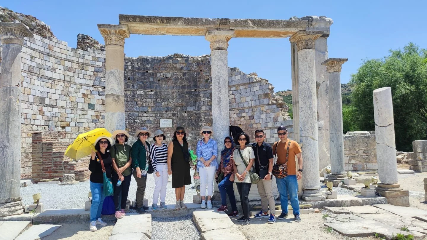 Group of tourists standing in front of ancient ruins with columns and stone walls, some wearing hats and sunglasses, under a clear blue sky.