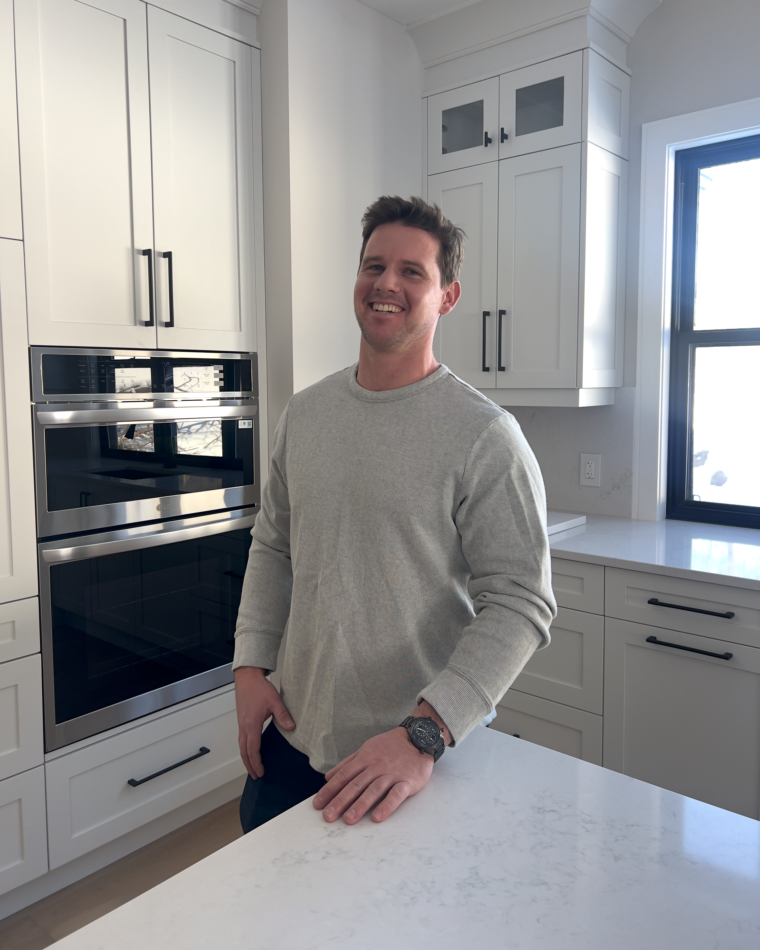 A smiling man standing in a modern kitchen with white cabinets, a black window, and a stainless steel oven.
