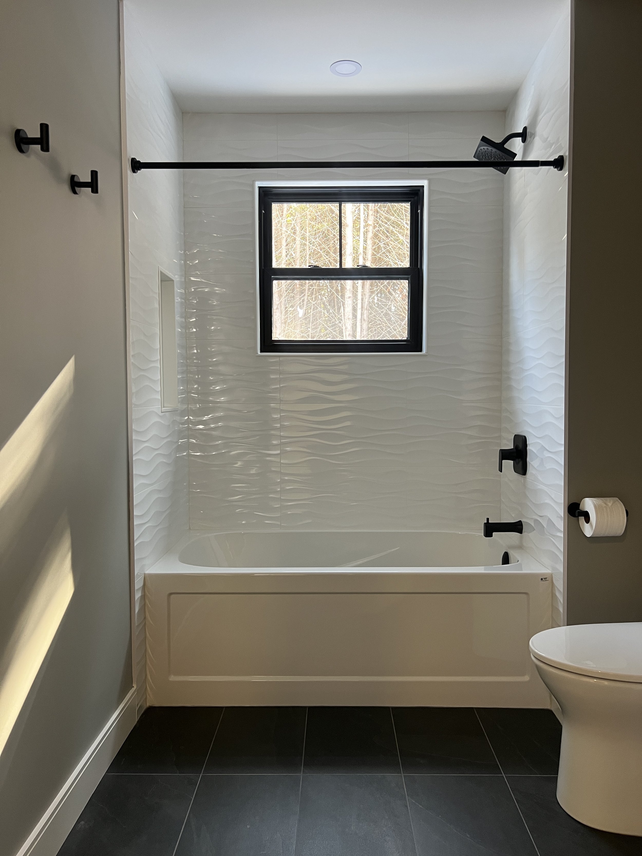 Modern bathroom with white tiled bathtub area, black window frame, black fixtures, and dark floor tiles.