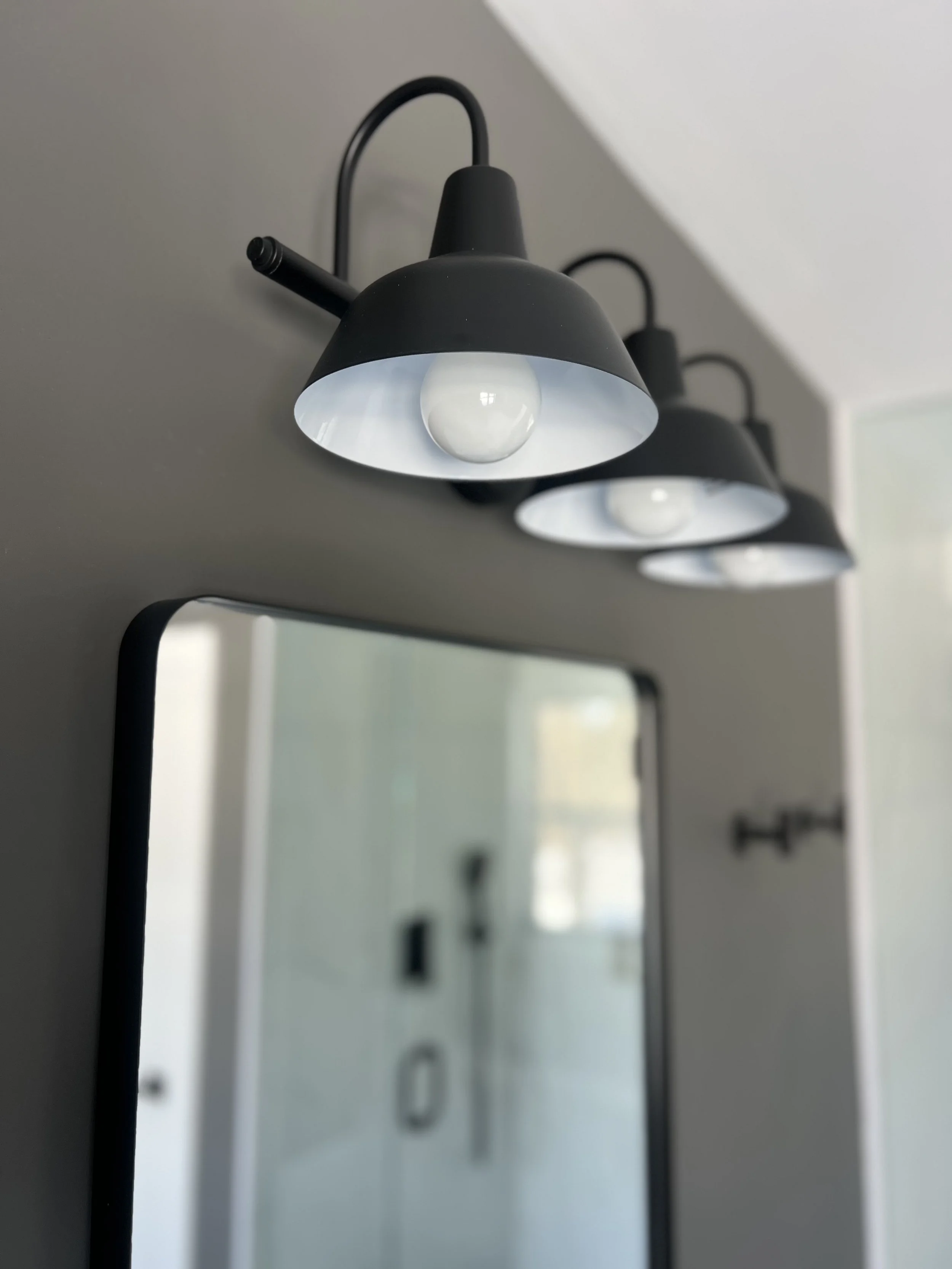 Close-up of three black, modern wall-mounted lamps with white interior, hanging above a mirror in a bathroom or hallway, with ceiling and part of a wall visible.