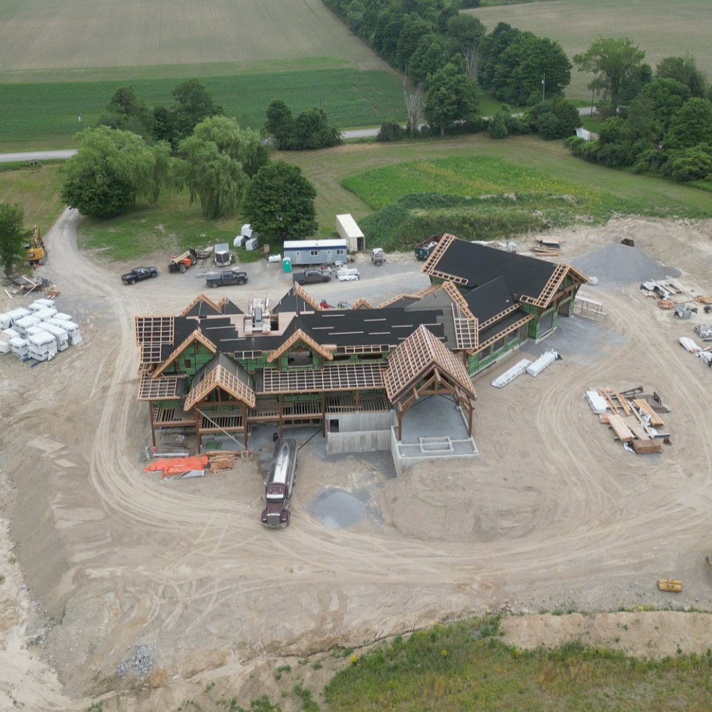 Aerial view of a large timber frame house under construction with exposed roof framing, surrounded by construction equipment, vehicles, and dirt grounds, with green fields and trees in the background.