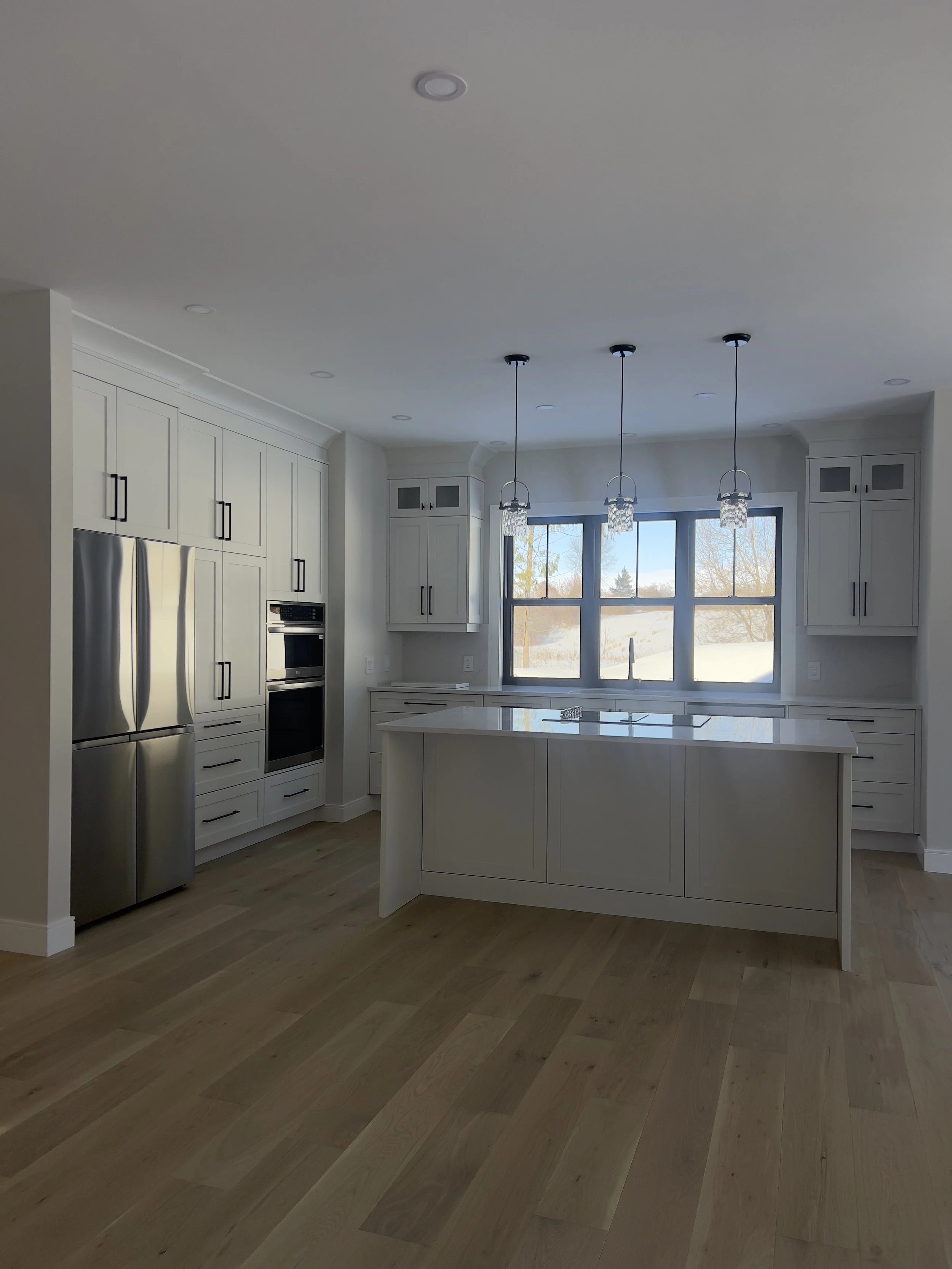 Spacious, modern kitchen with white cabinets, stainless steel refrigerator, built-in ovens, a kitchen island, large window, and hanging pendant lights.