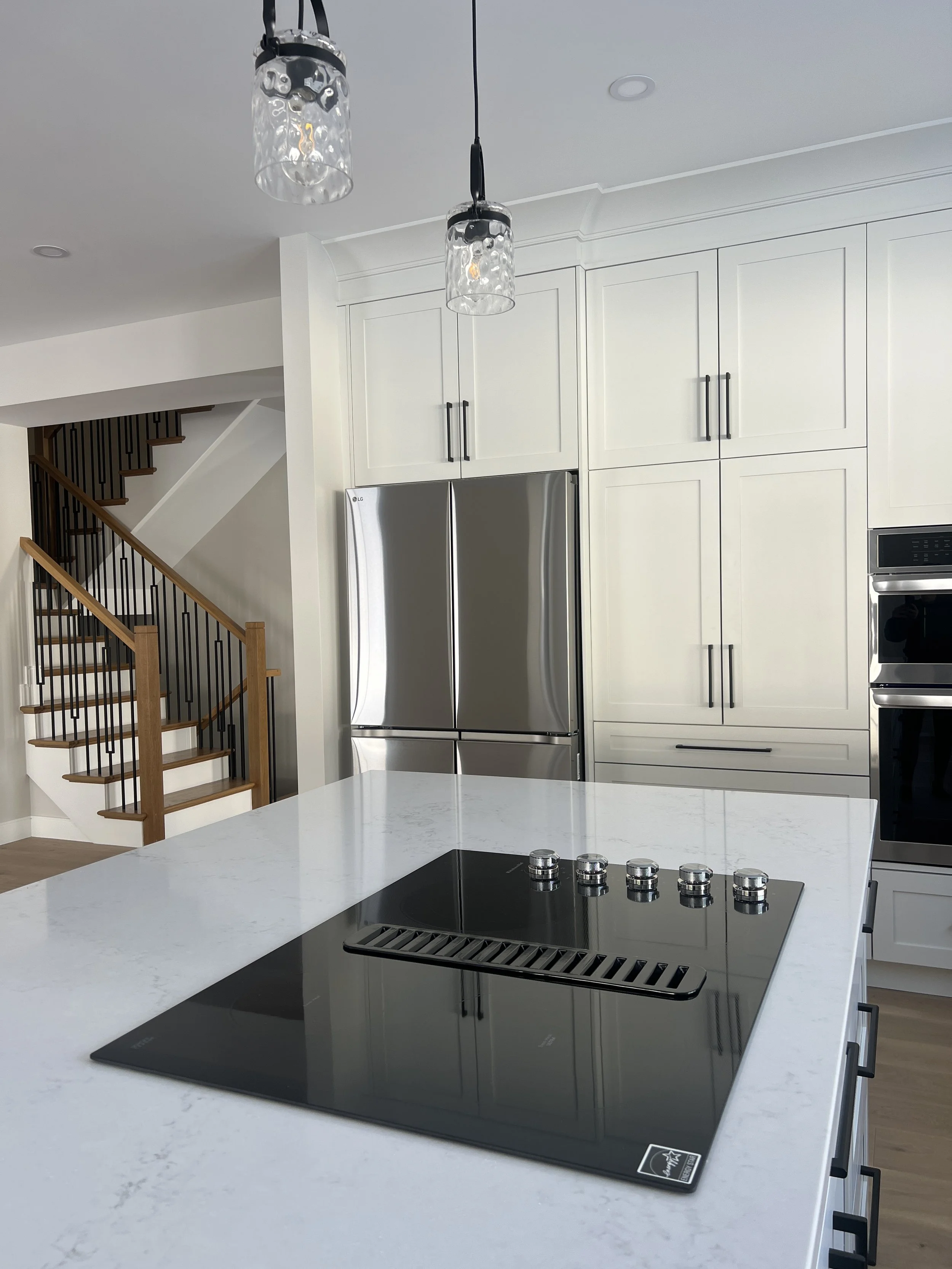 Modern kitchen with white marble countertop, stainless steel refrigerator, black glass electric cooktop, white cabinets, pendant lights, and wooden staircase in the background.