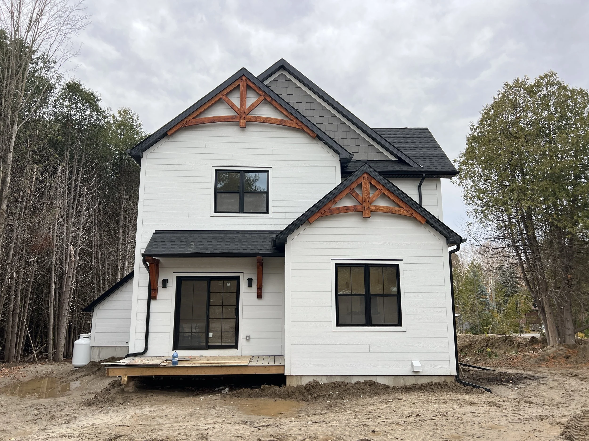 Newly built white modern farmhouse with black trim and decorative wooden gables with stain finish, surrounded by trees.