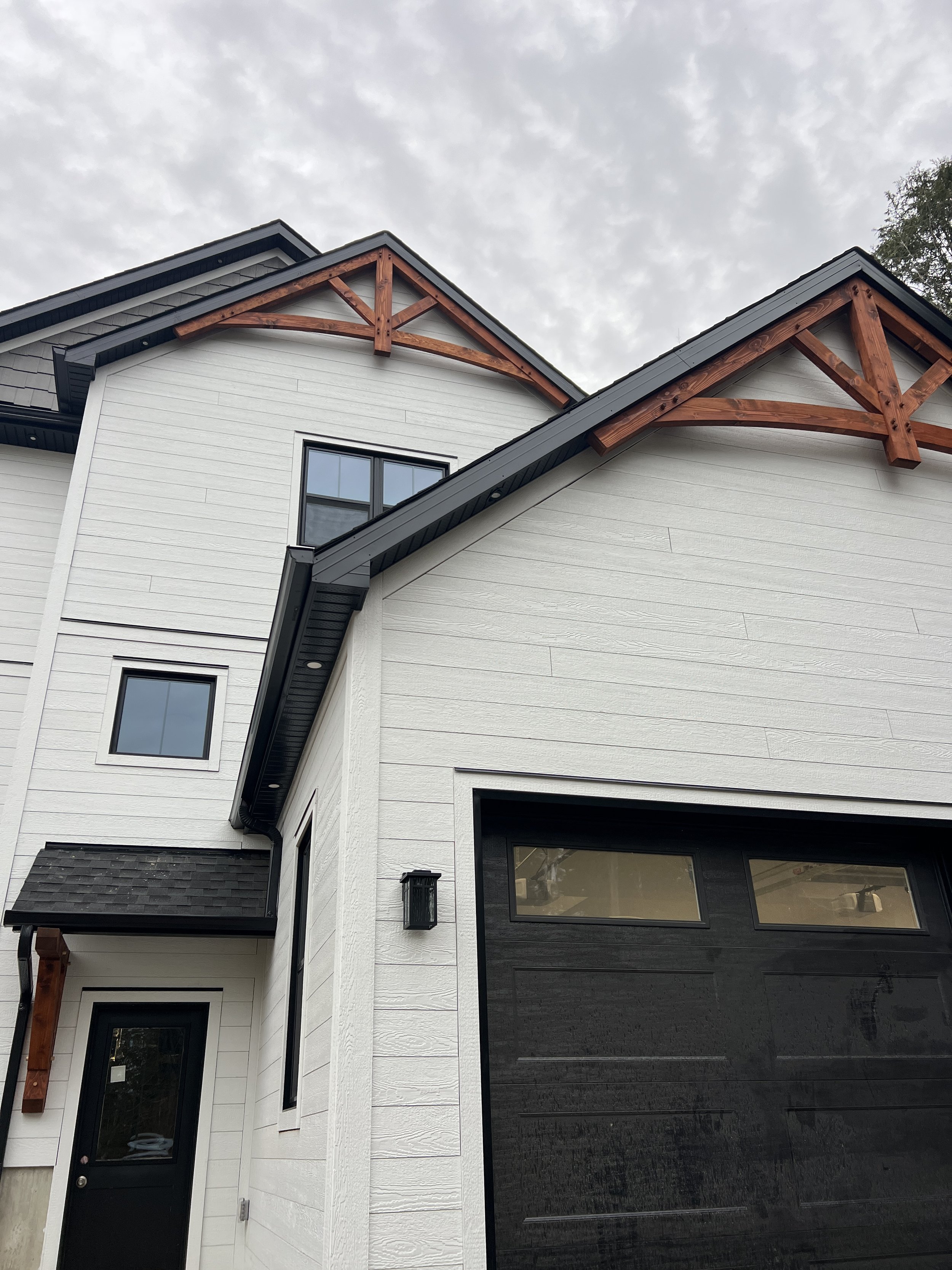 Close-up of a white modern farmhouse with black accents, featuring a garage door, black window frames, outdoor wall light, and decorative wooden gables with stain finish.