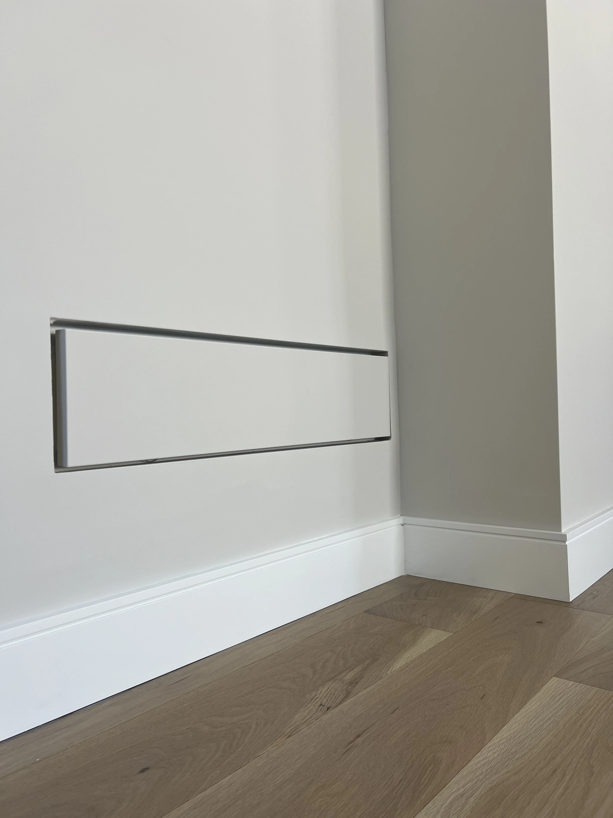 Close-up of a white wall vent near the baseboard on light wood flooring.