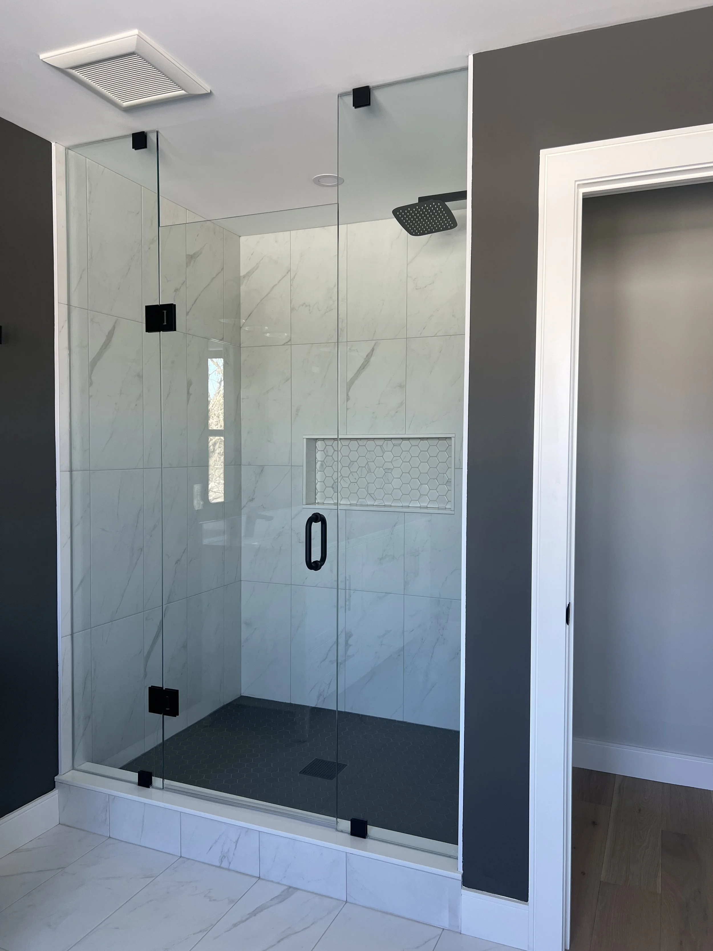 A modern walk-in shower with frameless glass doors, white marble tiled walls, a black rainfall showerhead, and a built-in niche with hexagonal tiles, inside a contemporary bathroom with light-colored tile flooring and dark gray walls.